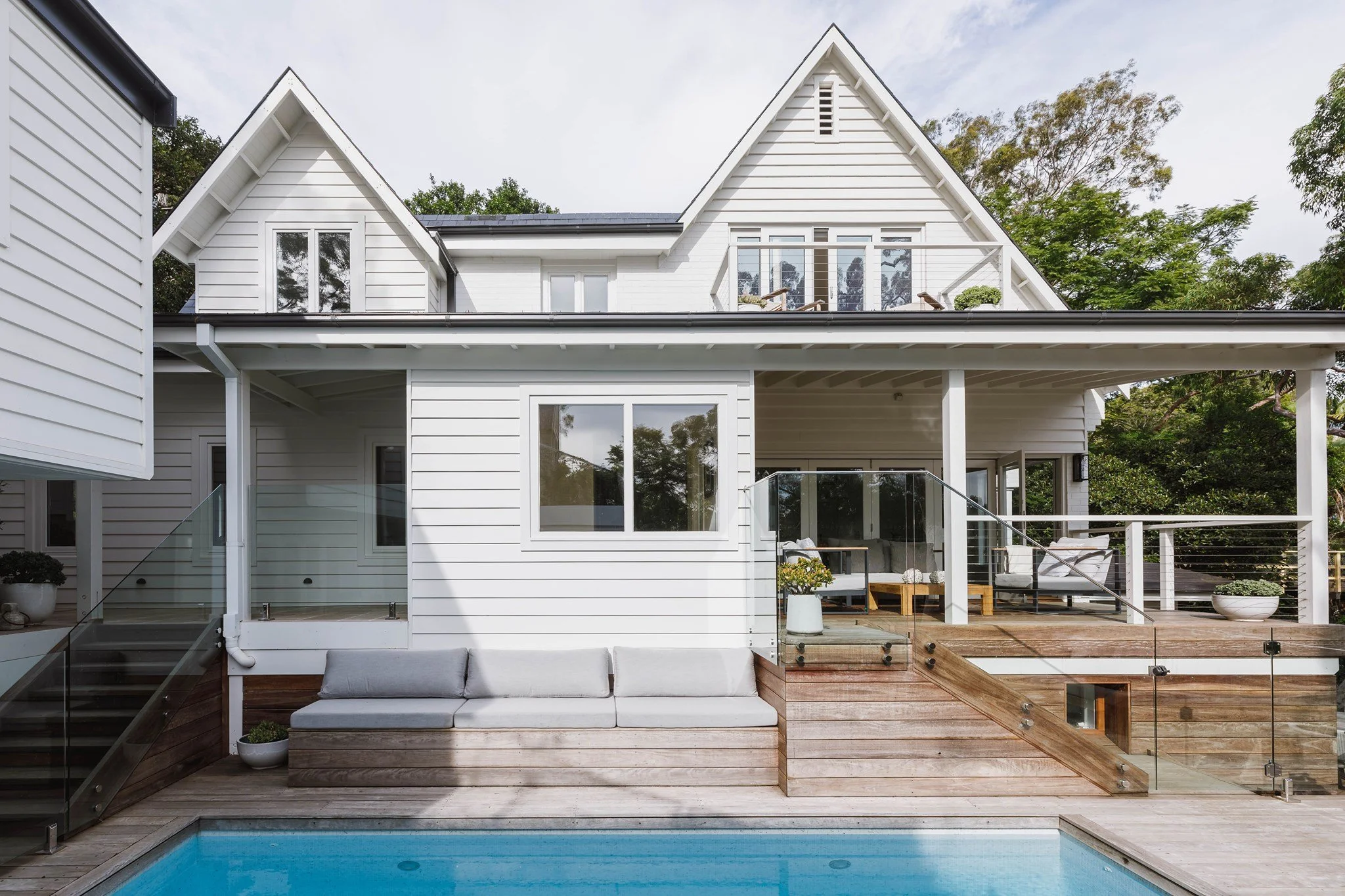 Modern white house with a wooden deck and pool, featuring large windows and glass railings, surrounded by trees.