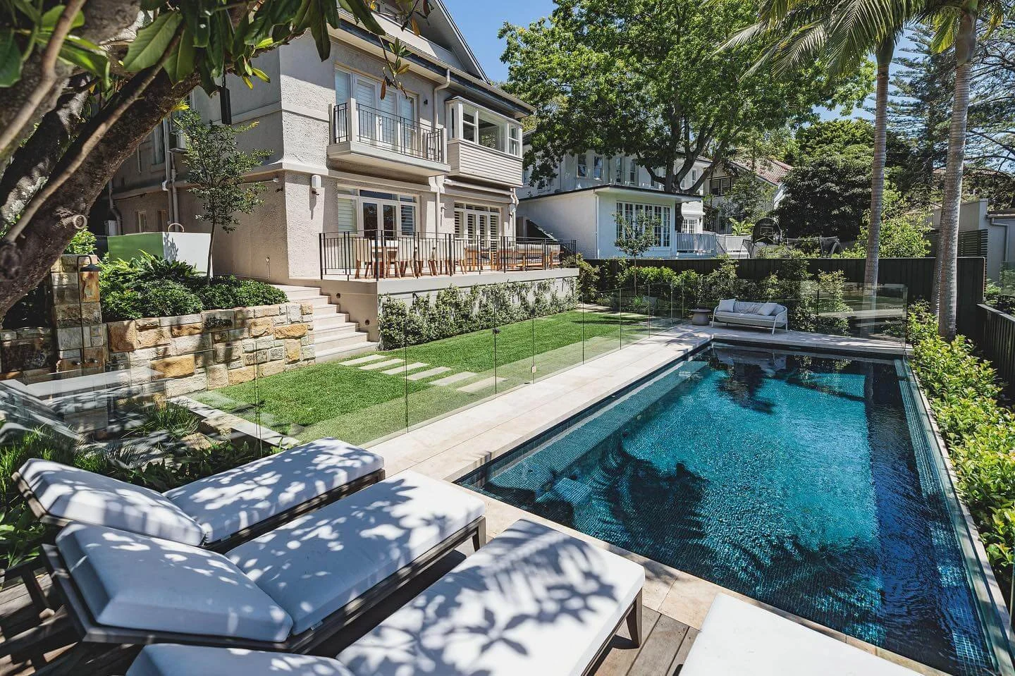Backyard with a swimming pool, lounge chairs, a grassy area with stepping stones, and a multi-story house with a balcony and patio furniture, surrounded by trees and greenery