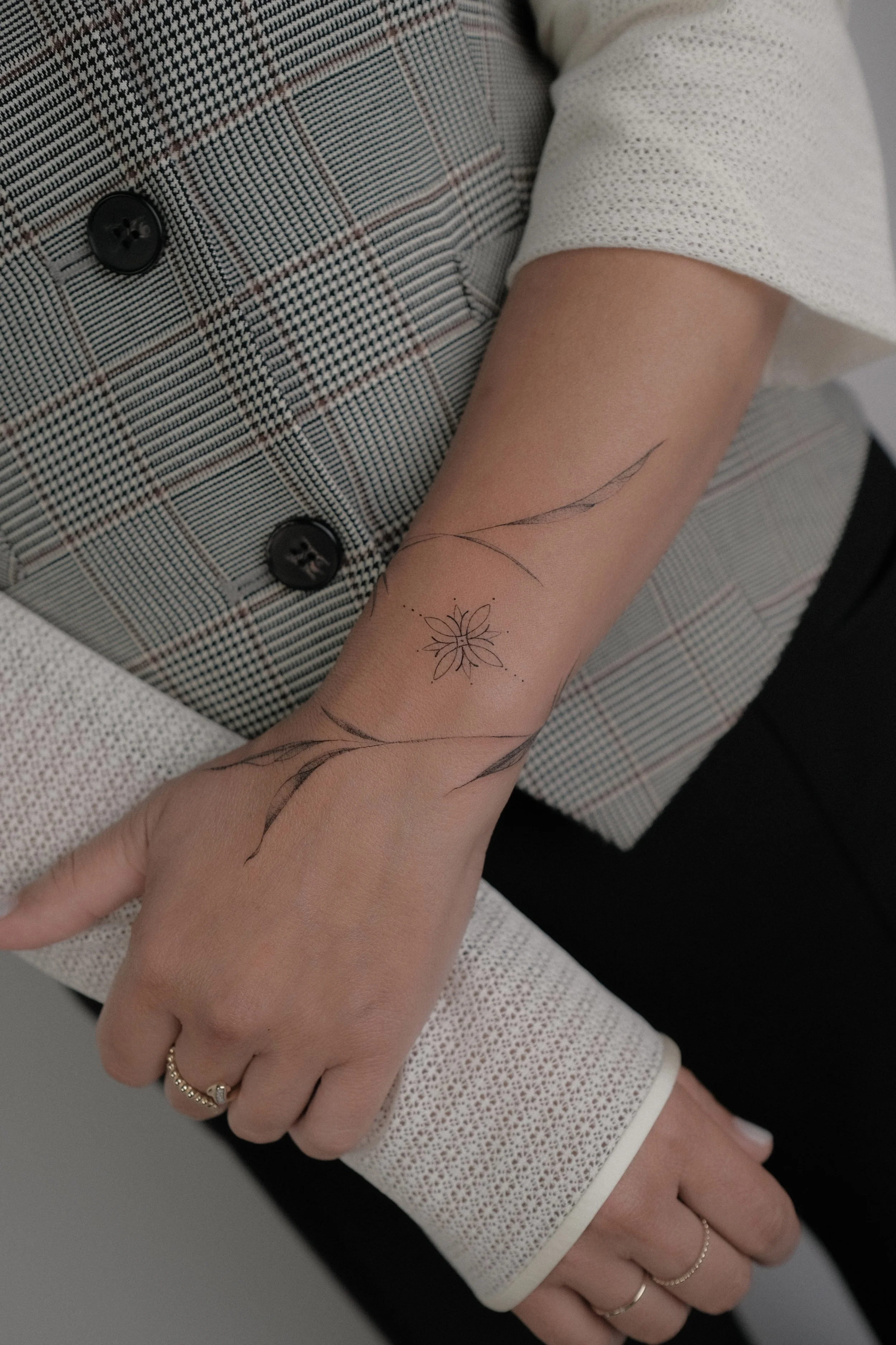 A person's arm showing a delicate floral tattoo with leaves and a small flower, with some dotted lines on the skin, and wearing rings on fingers. The person wears a buttoned, checkered blazer with a white sleeve underneath.