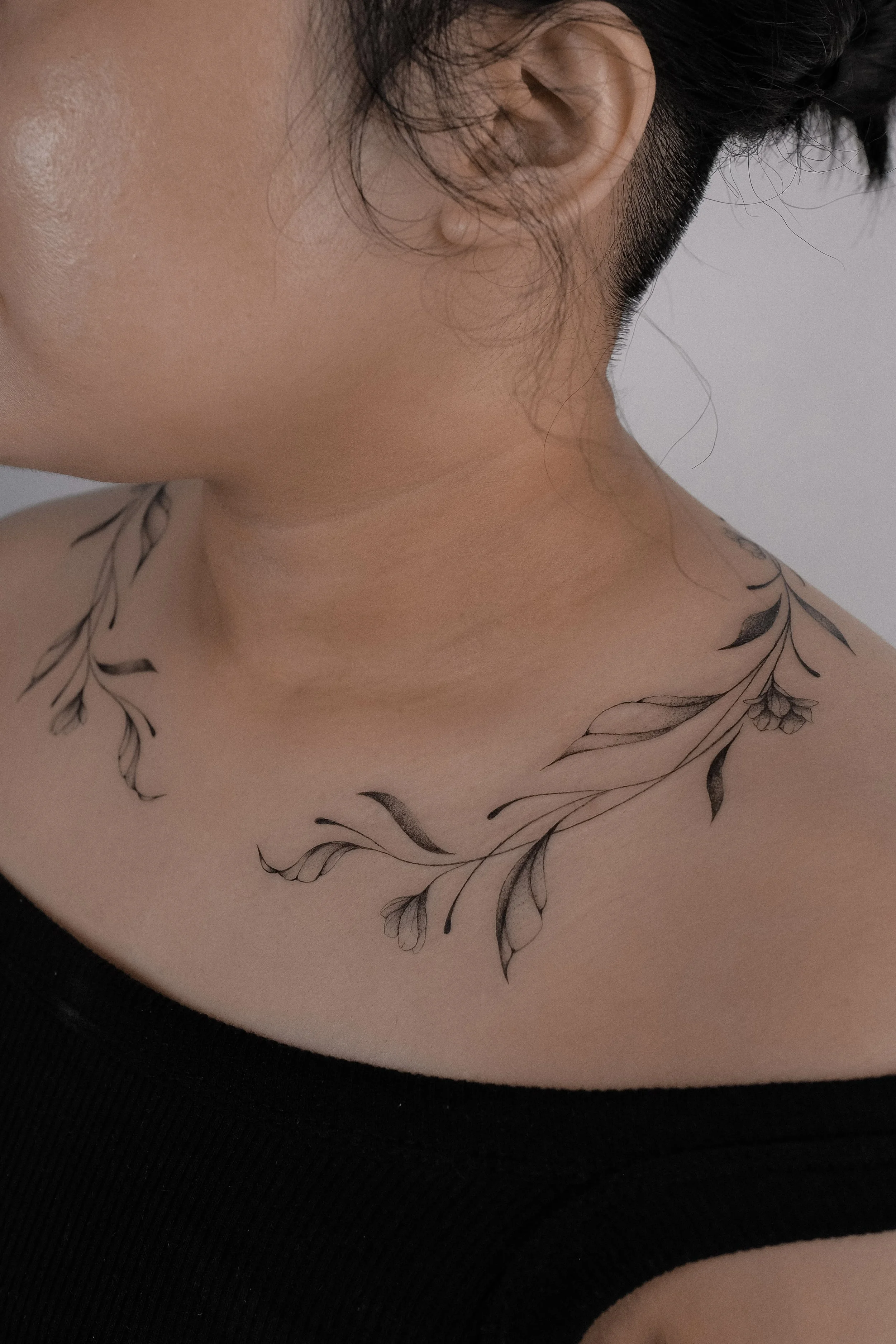 Close-up of a person's neck and shoulder with a floral tattoo, wearing a black top, and with part of their face and ear visible.