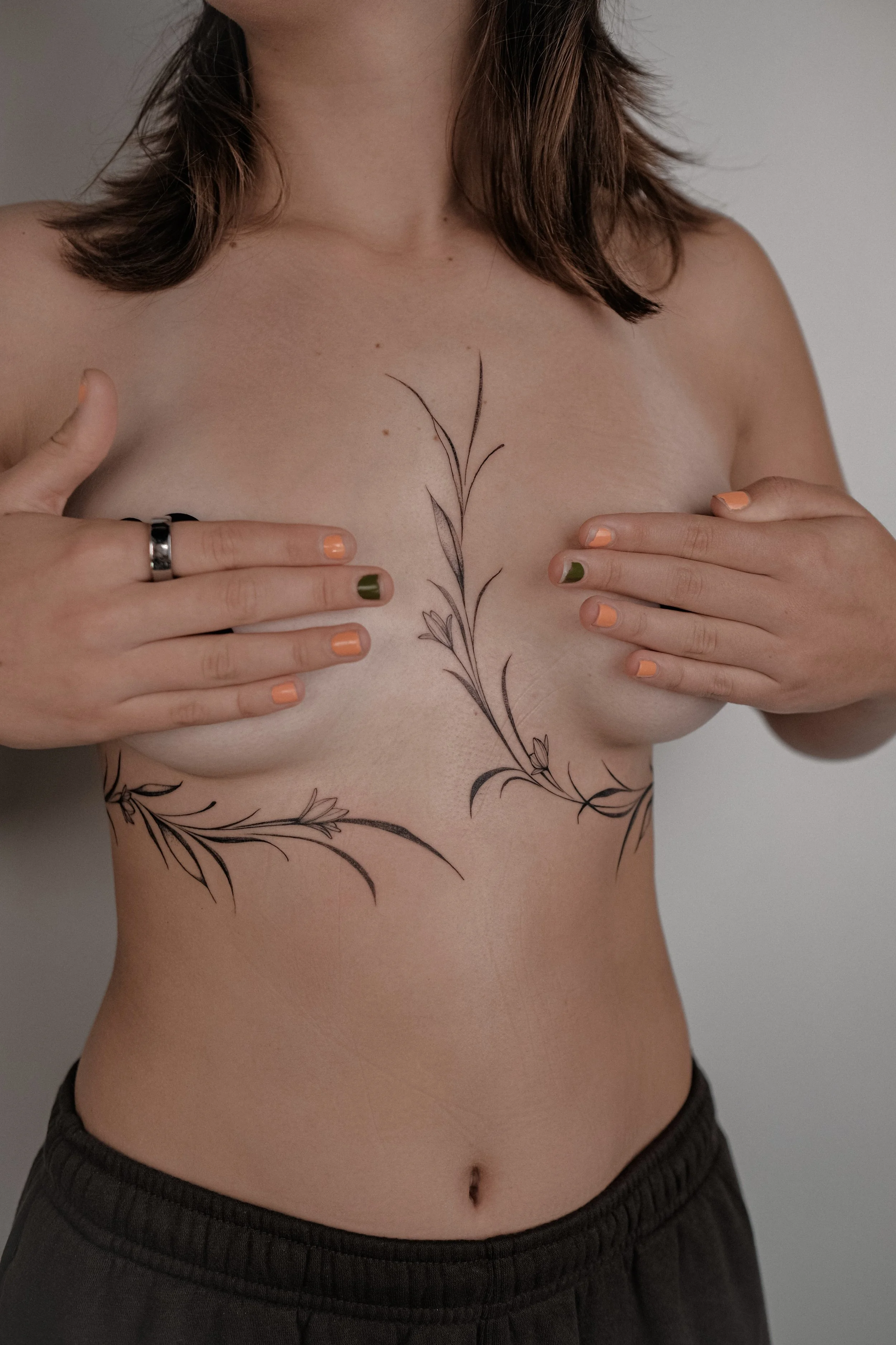 A woman covering her breasts with her hands, featuring a large floral tattoo across her chest and sides.