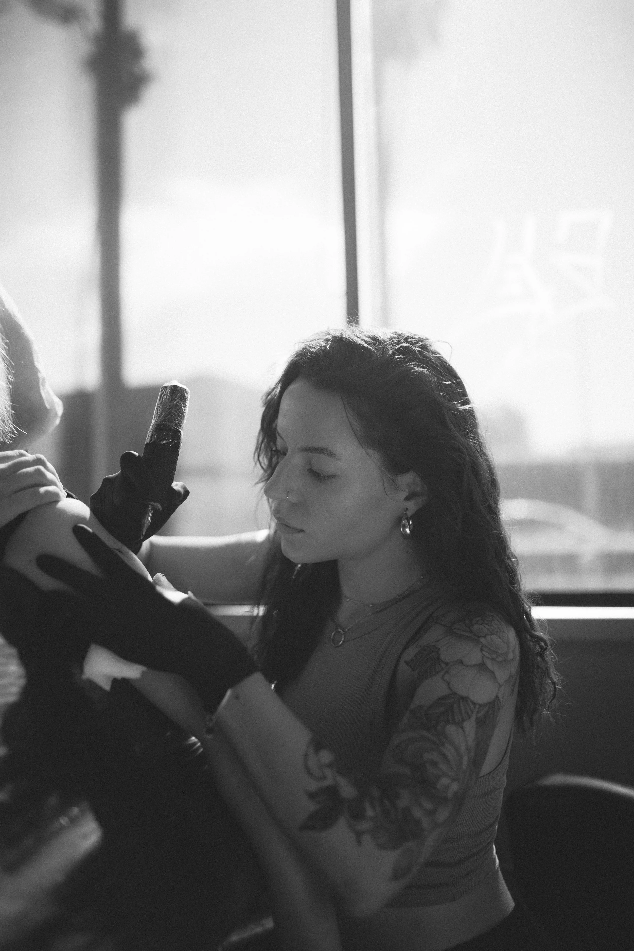 A young woman with dark wavy hair, tattoos on her arm, and earrings, is getting a tattoo with her eyes closed. A tattoo artist is working on her arm.