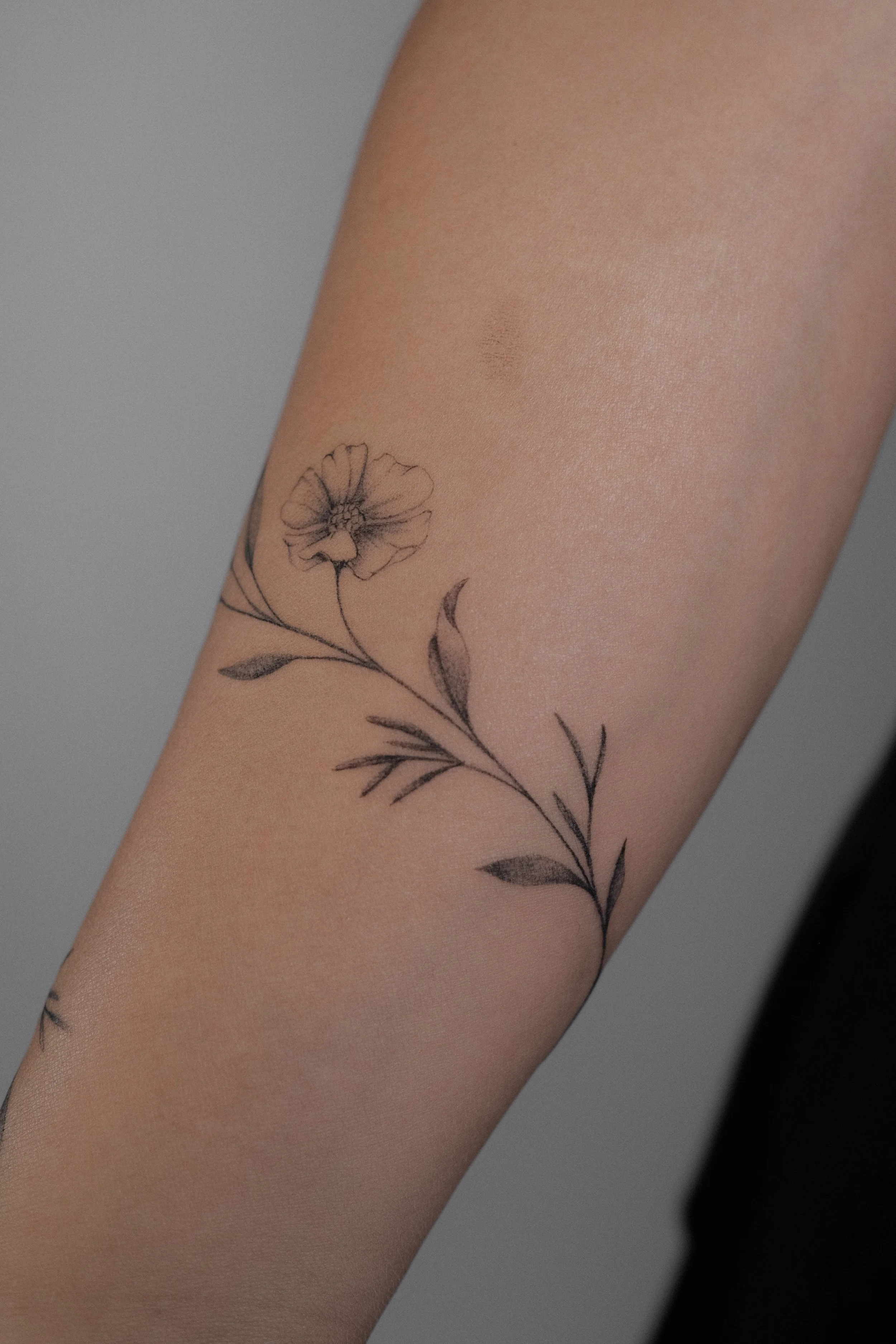 A tattoo of a delicate black outline flower with leaves on a person's forearm.