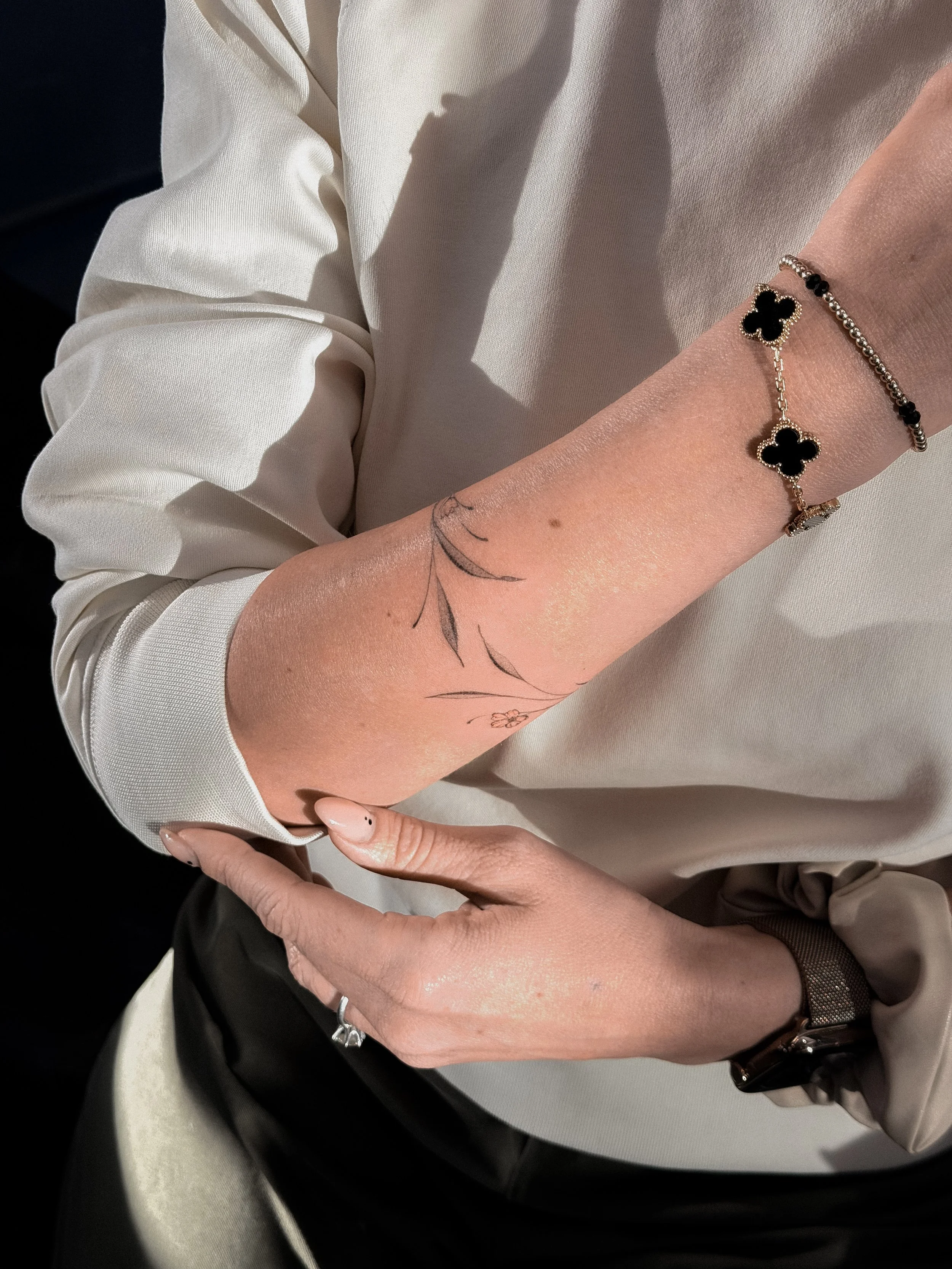 A person's arm with a tattoo of a plant stem and small flower, wearing a gold bracelet with black and gold four-leaf clovers, a black beaded bracelet, and an Apple Watch. The person is dressed in a light-colored long-sleeve shirt.