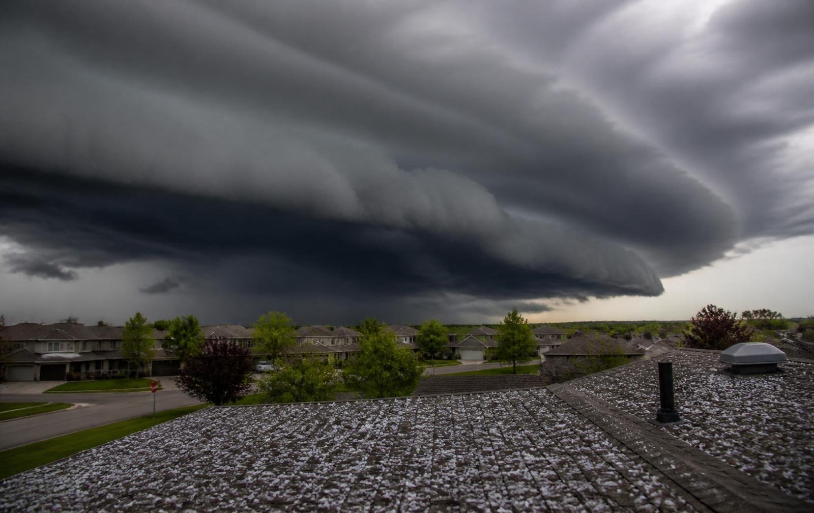 Dark and ominous storm clouds looming over a suburban neighborhood, with rain pouring on rooftops and green trees on a cloudy day.