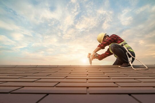 A construction worker installing shingles on a roof during sunset, wearing a safety helmet and vest.