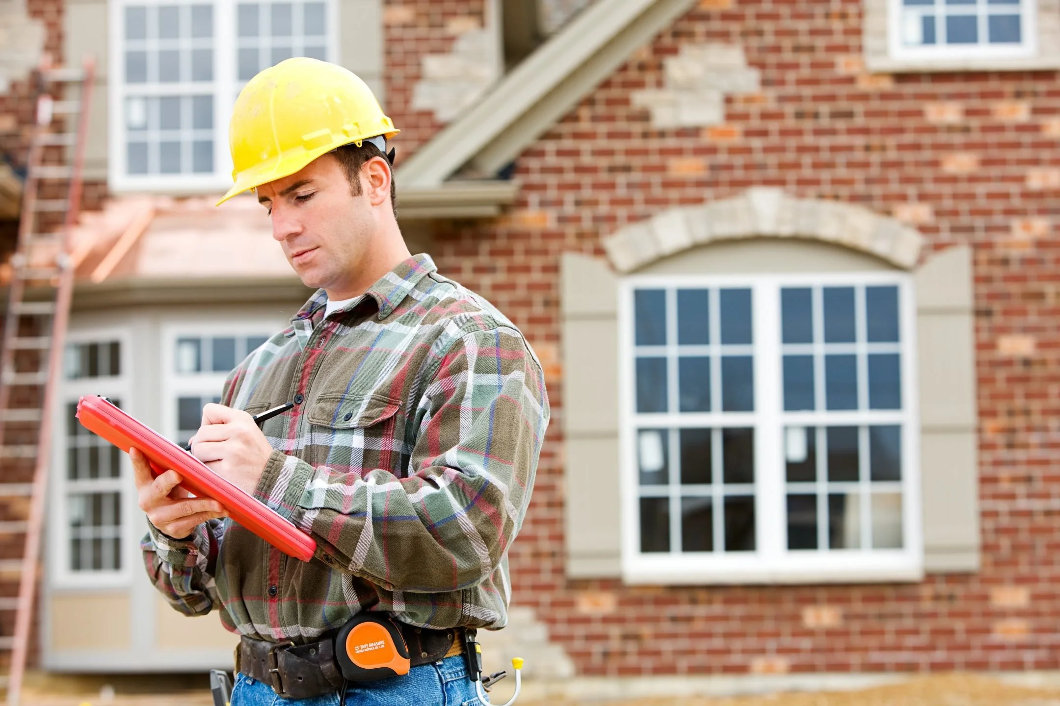 A construction worker wearing a yellow safety helmet and plaid shirt, holding a tablet, on a house construction site with a brick house and scaffolding in the background.