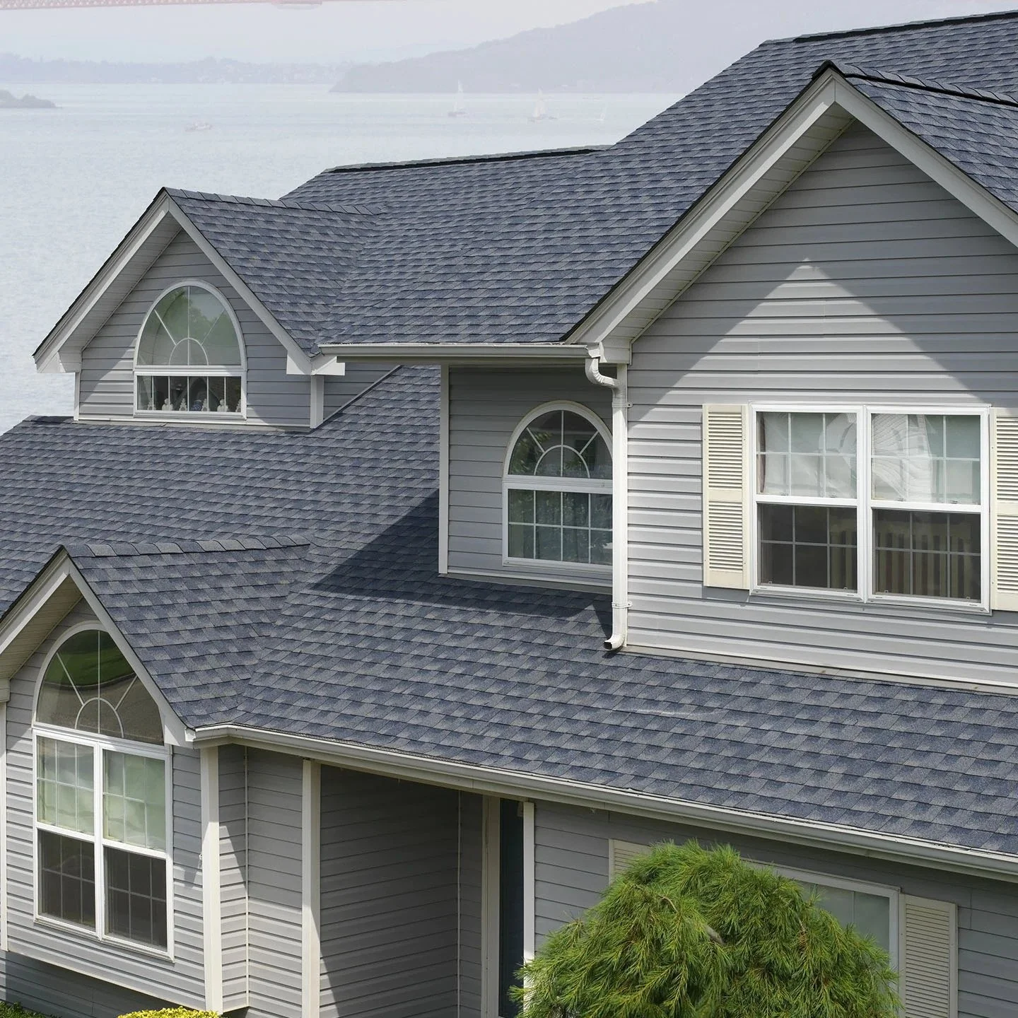 A multi-story house with gray siding, multiple large arched and rectangular windows, and a blue shingle roof, located near a body of water with sailboats in the background.