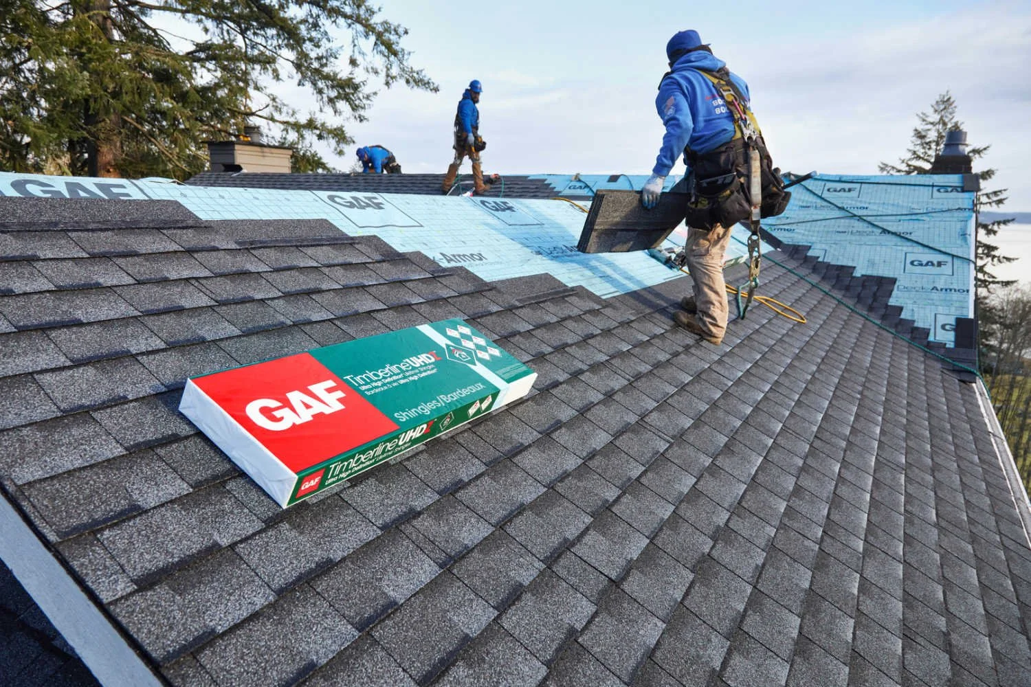 Workers installing asphalt shingles on a residential roof with GAF Timberline UHD shingle bundle visible in the foreground.