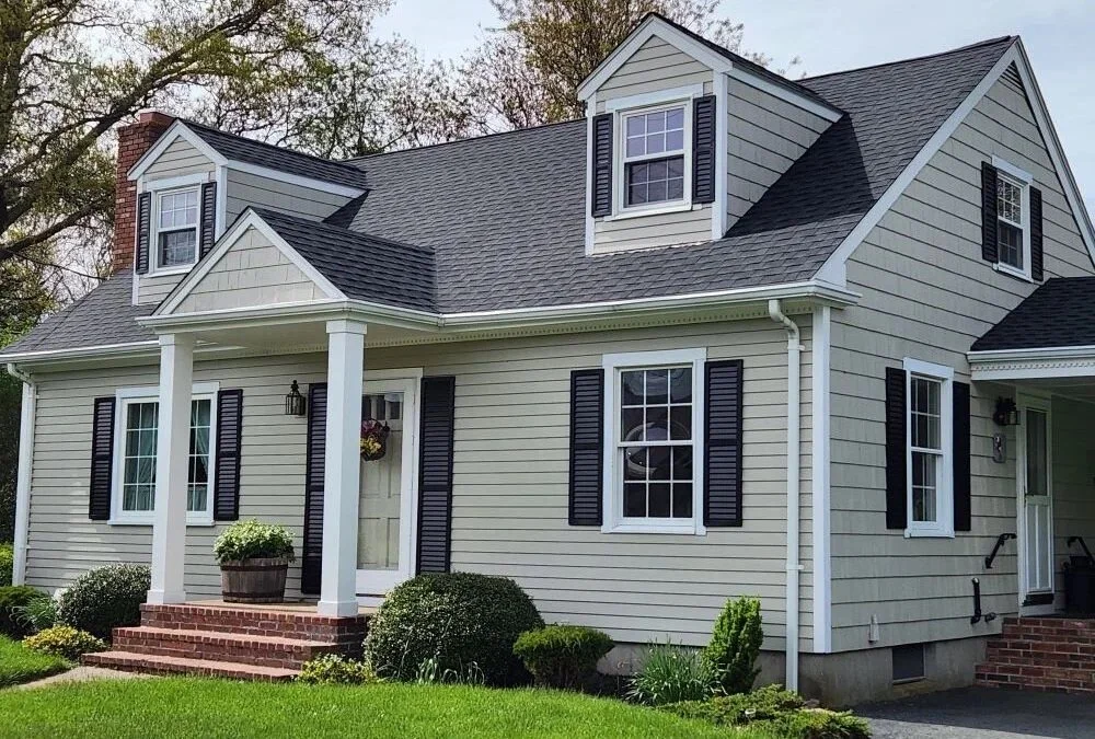 A two-story house with a beige exterior, black shutters, white trim, and a gabled roof. There are three dormer windows and a small front porch with brick steps, white columns, and a potted plant.