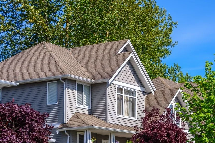 A two-story house with gray siding and a brown shingled roof, surrounded by green trees and bushes under a clear blue sky.