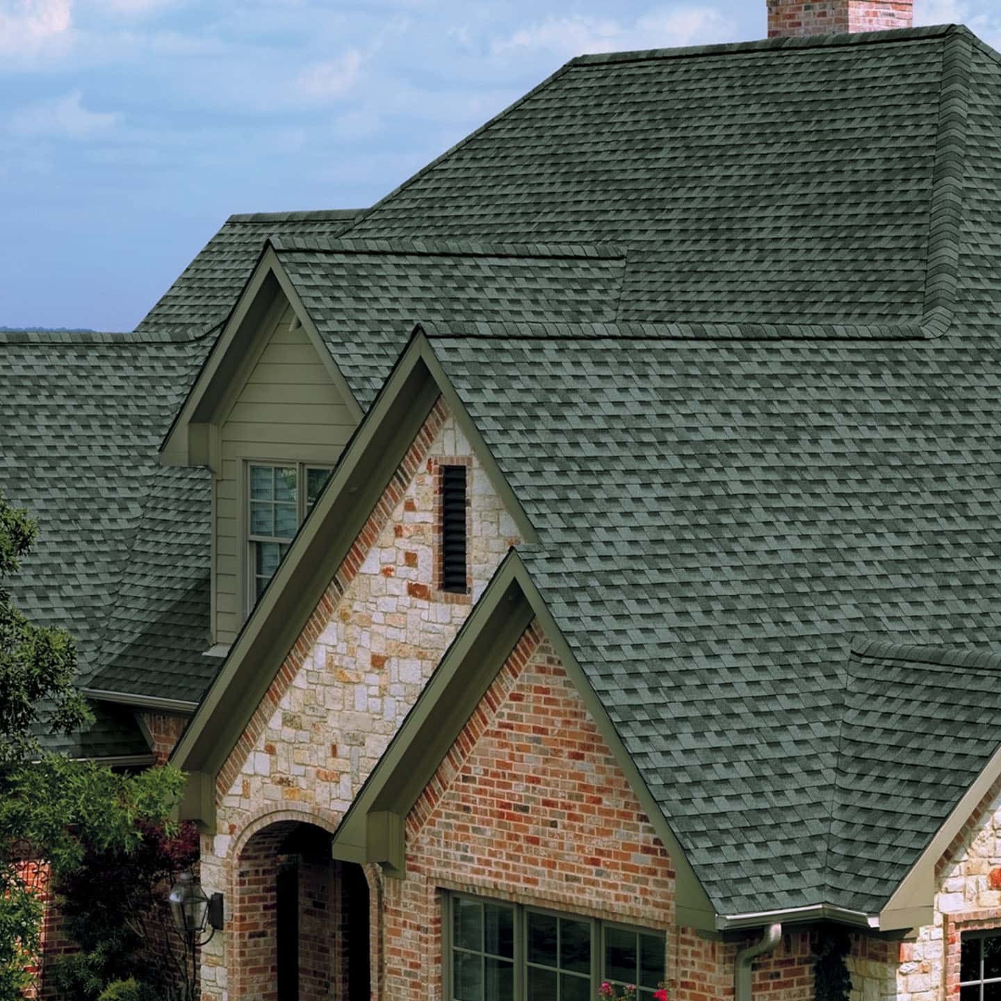 Close-up of a house's roof featuring green shingles, brick and stone exterior walls, and a small window with a black vent beneath it.