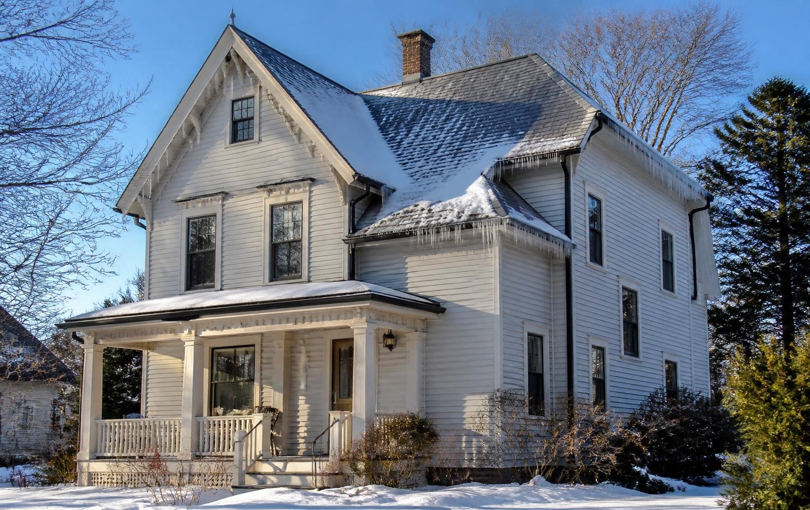 A large, white, three-story house with a porch, black window frames, and a steep roof with icicles hanging from the eaves, surrounded by snow and leafless trees.
