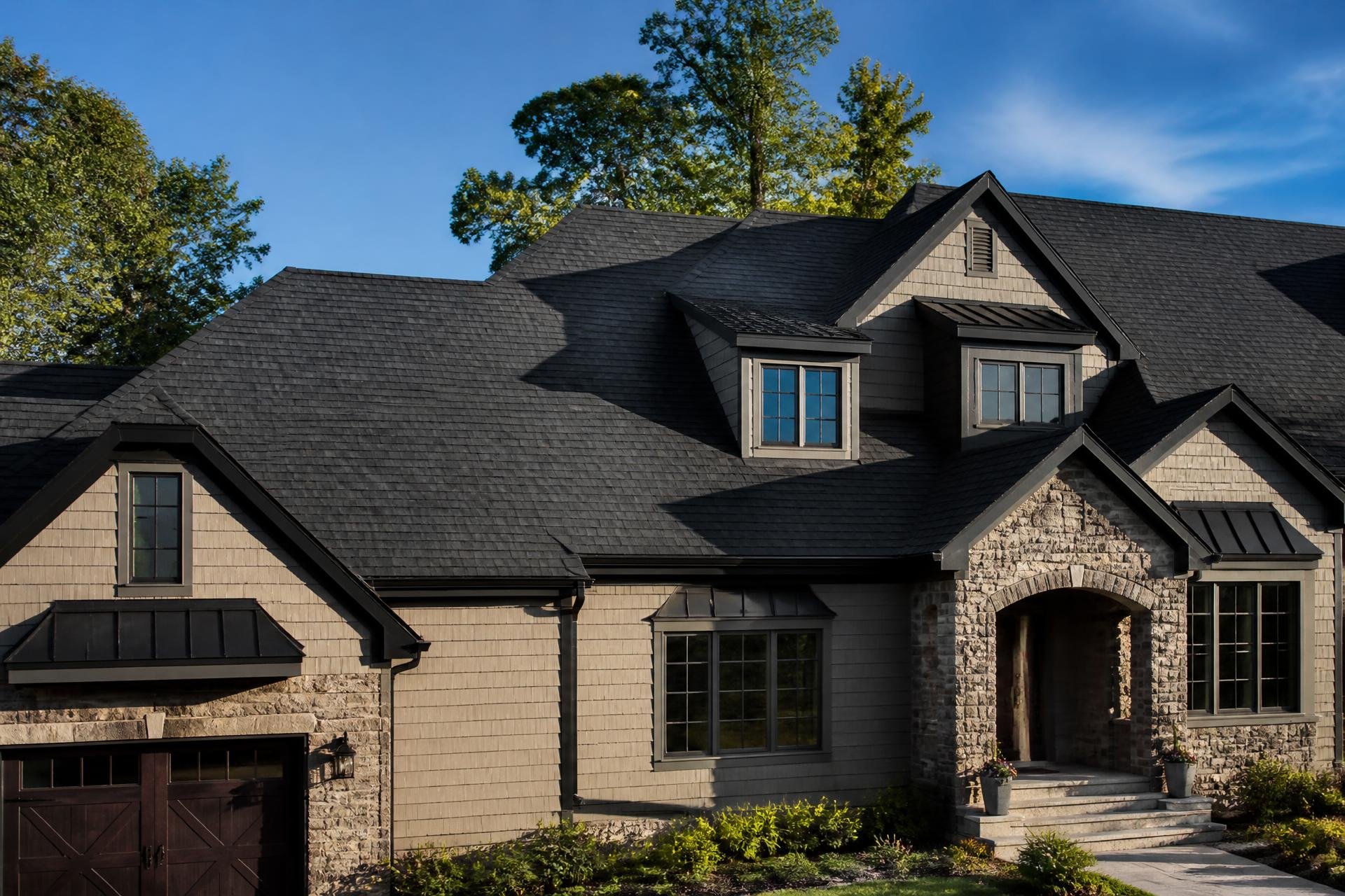 A two-story house with a combination of stone and beige siding, multiple gabled roofs with black shingles, and several windows. The house has front steps leading to a covered entrance with potted plants on either side, surrounded by a green lawn and trees in the background.