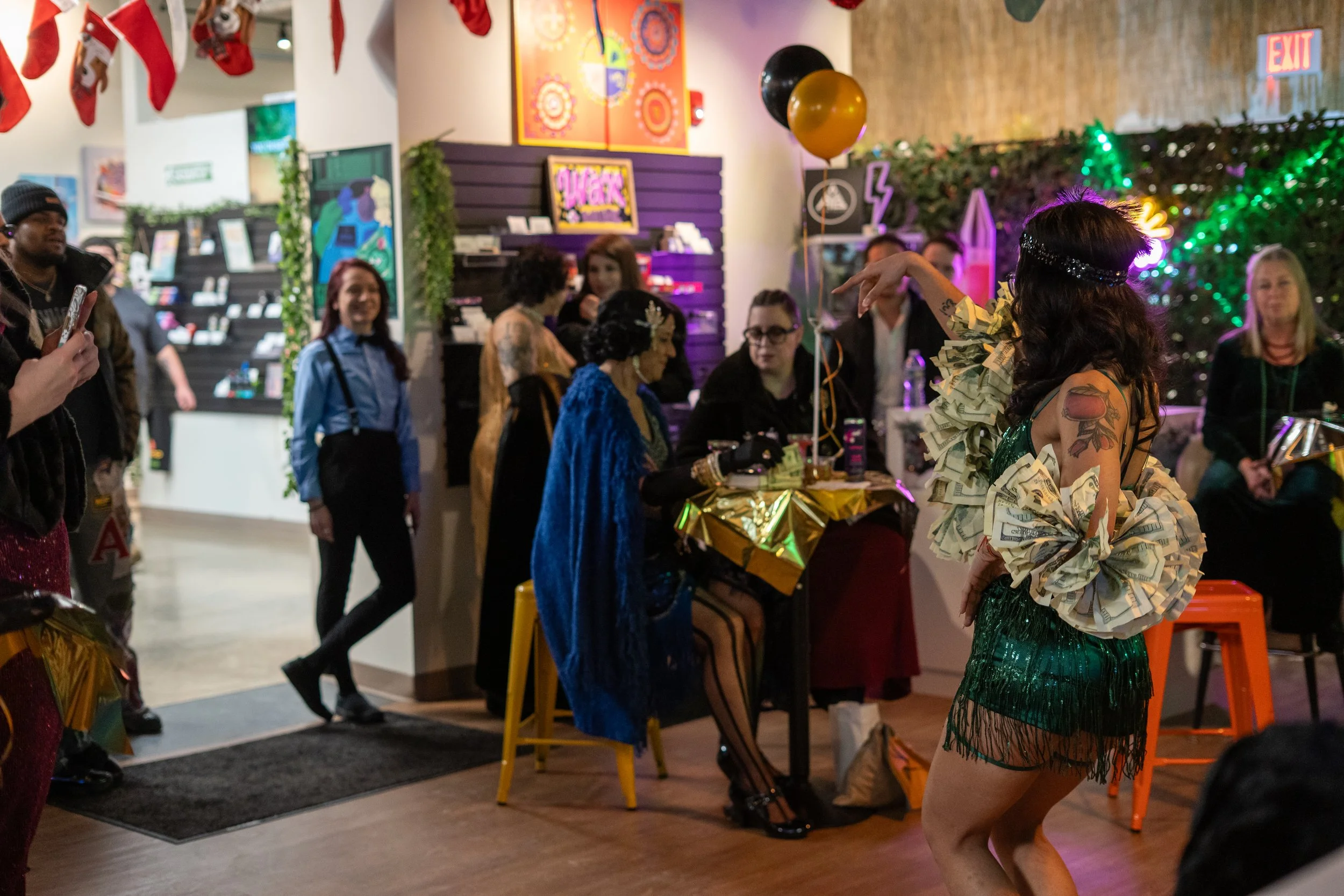 A woman with tattoos performing a dance in a festive indoor setting decorated with balloons, Christmas stockings, and colorful lights, while an audience of diverse people watch and take photos.