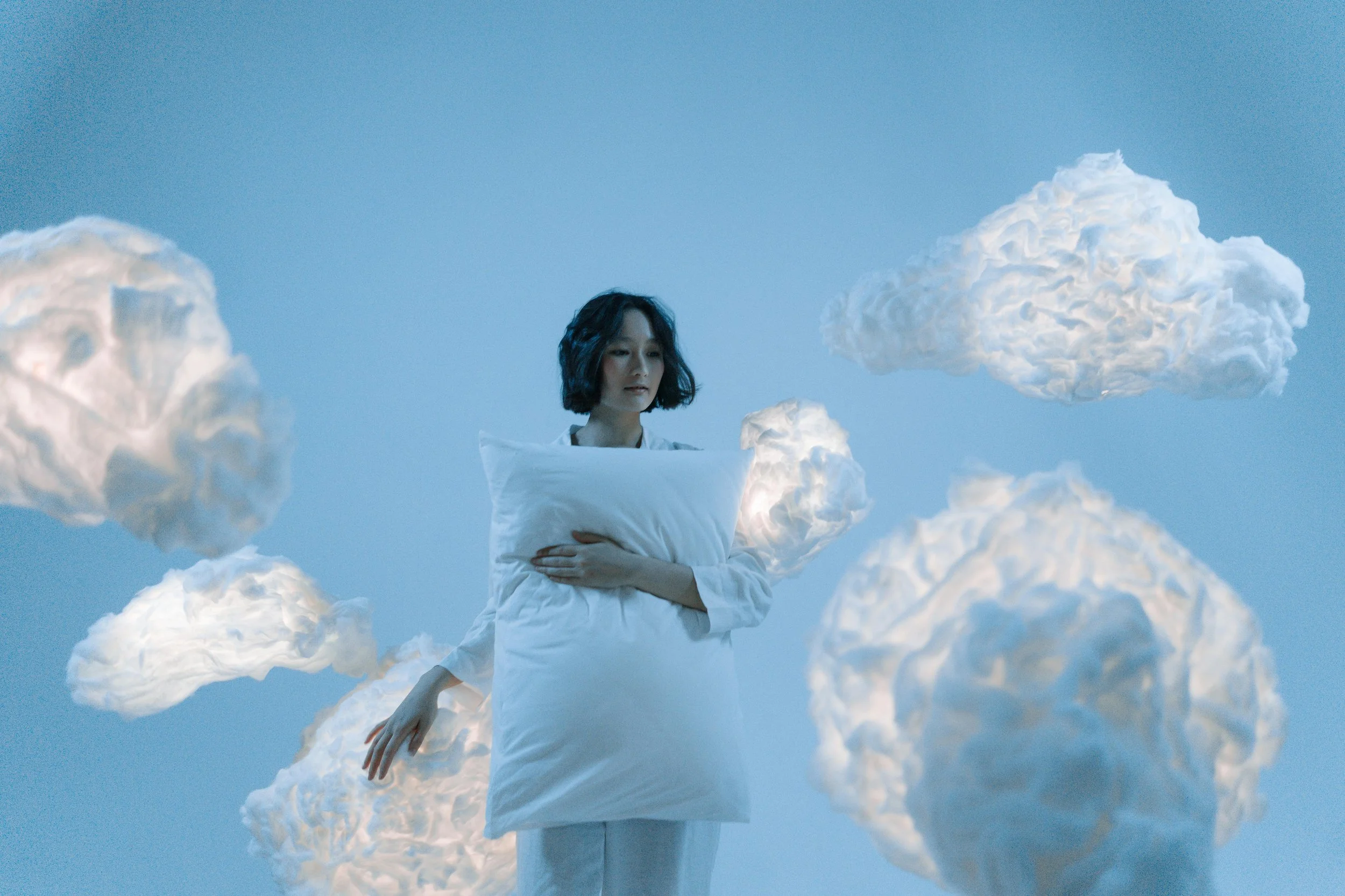 A woman with black wavy hair wearing a white dress, standing among white clouds against a blue sky background.
