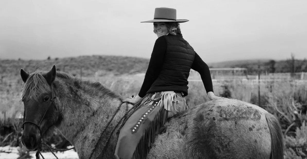 A woman smiling and riding a horse in an outdoor rural landscape, wearing a wide-brimmed hat and a black vest.