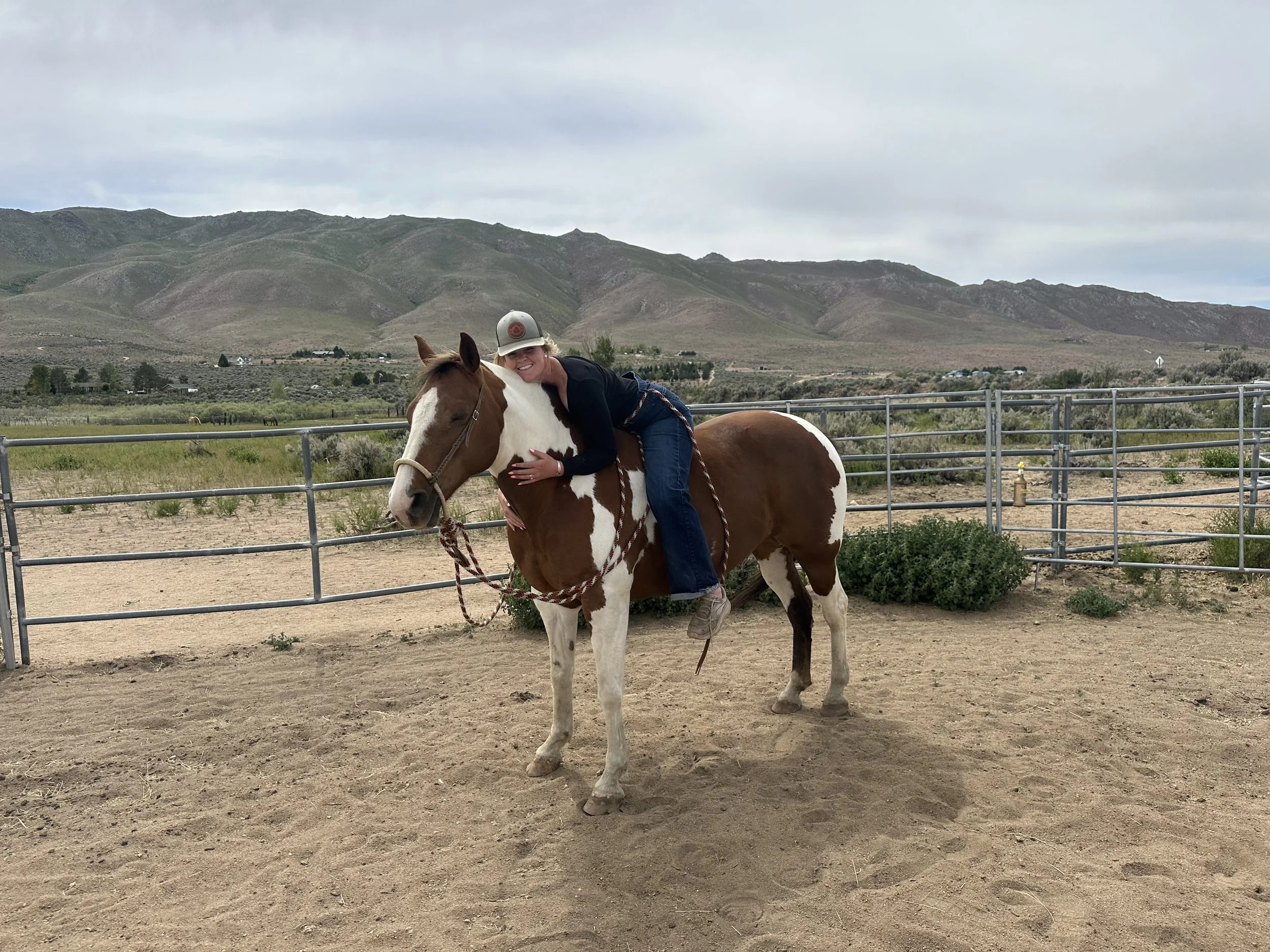 Woman hugging a brown and white pinto horse in a fenced outdoor arena with mountainous landscape and cloudy sky in the background