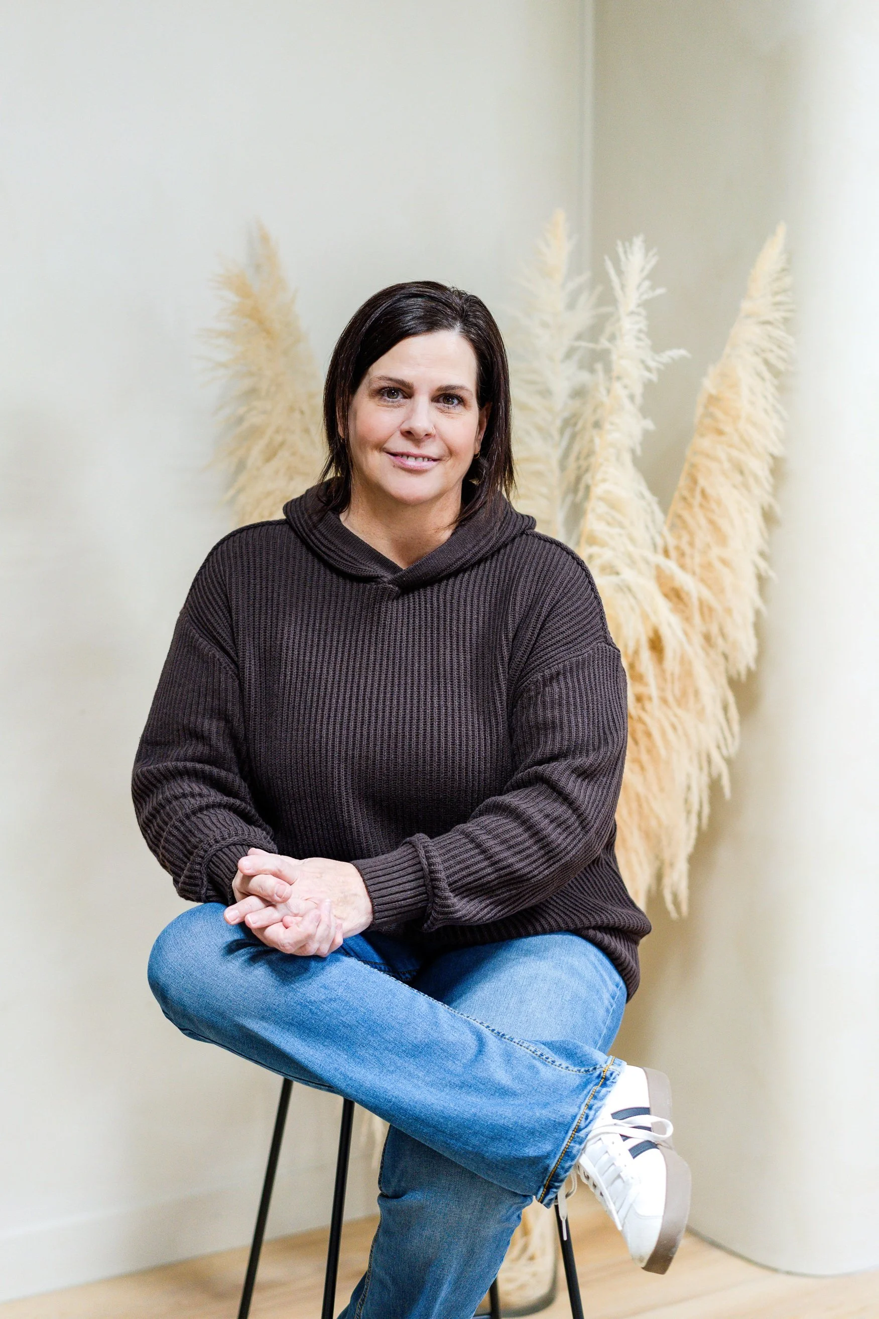 A woman with dark hair sitting on a black chair against a cream wall with tall dried pampas grass in the background, wearing a brown sweater and blue jeans, with crossed legs and her hands resting on her knee.