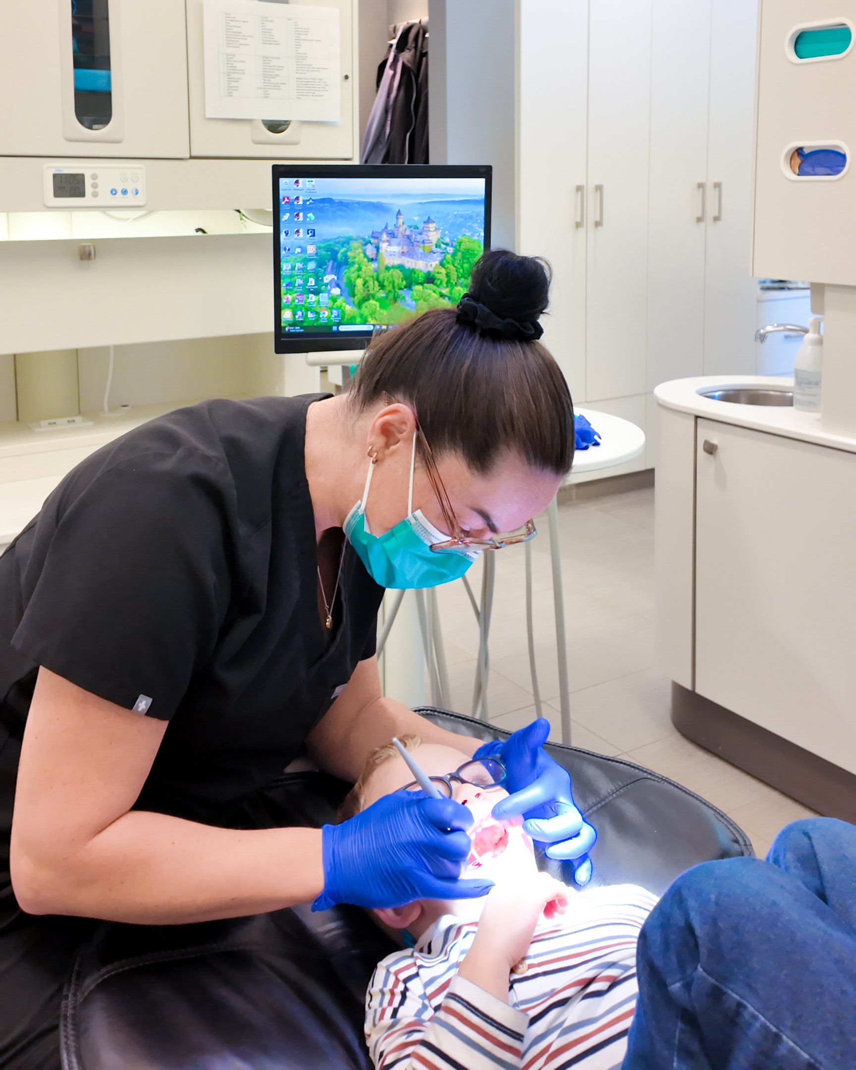 A dental professional wearing a face mask and blue gloves attends to a child's mouth during a dental procedure in a clinical setting with white cabinets and a computer monitor showing a castle and green landscape.