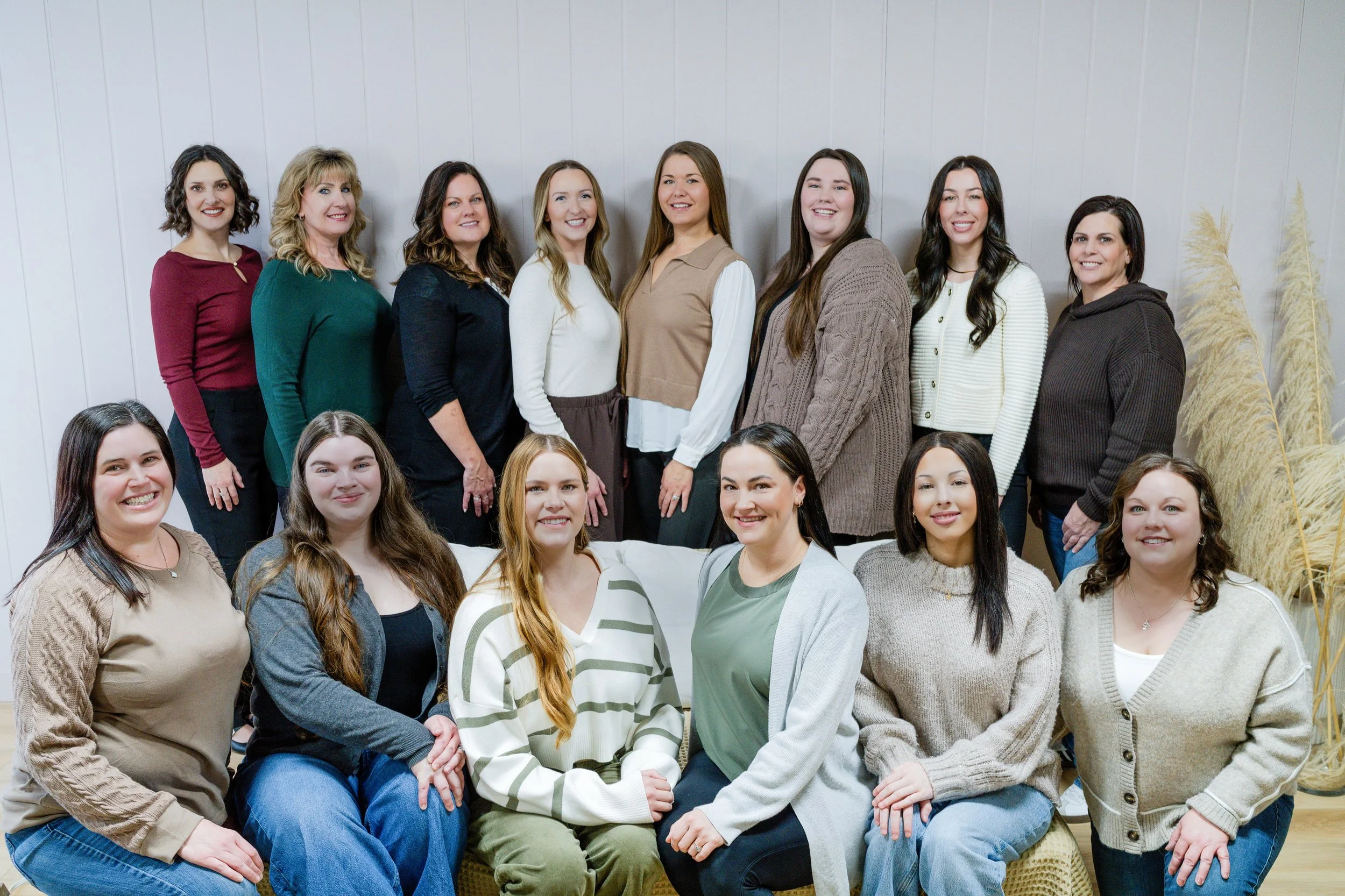 A group of women gathered together indoors, smiling at the camera, standing and sitting in front of a plain white wall with tall dried plants on the right side.