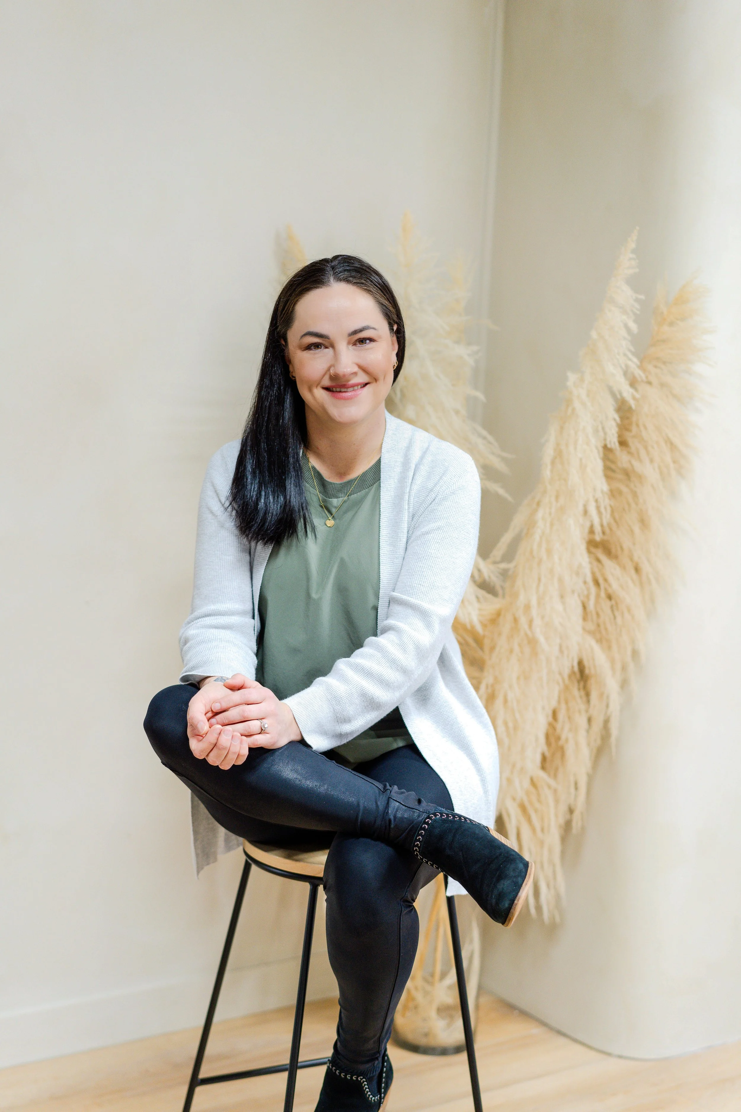 A woman with dark hair, smiling, sitting on a stool in a room with light-colored walls and beige pampas grass in the background. She is wearing a green top, a light cardigan, black pants, and boots.