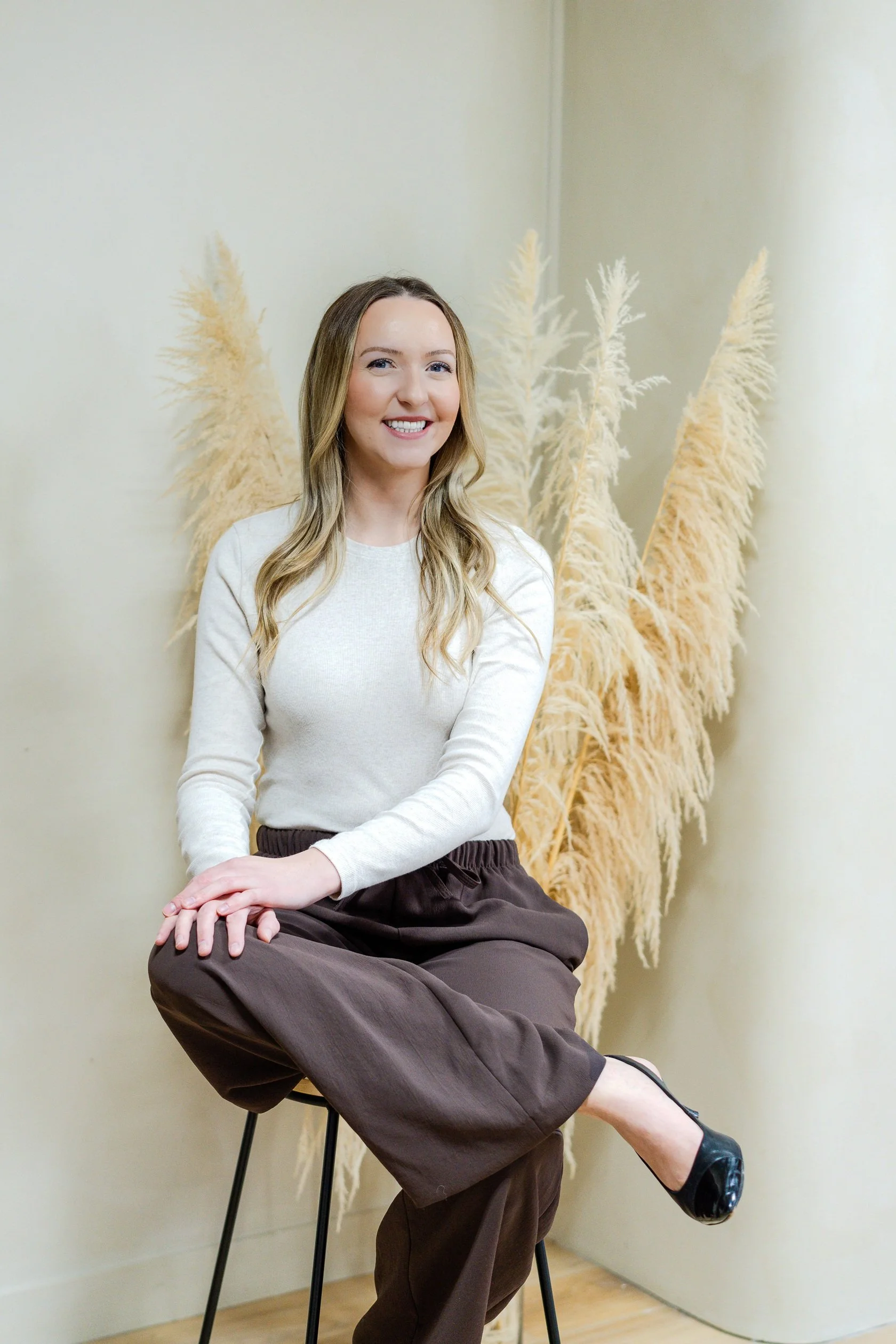 A woman sitting on a black chair with beige pampas grass behind her, wearing a white top, brown wide-leg pants, and black shoes, smiling and crossing her legs in a room with light-colored walls.
