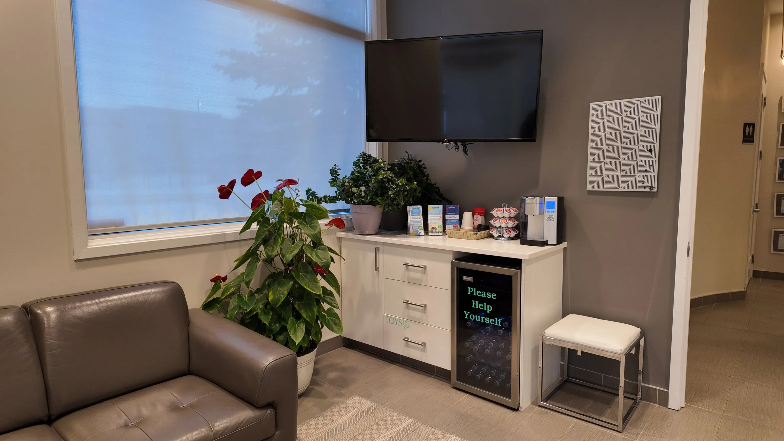 A lounge area with a large window covered by a translucent shade, a brown leather sofa, various potted plants, a white counter with informational flyers, cups, and a coffee machine, a small refrigerator with a digital sign that reads "Please Help Yourself," a wall-mounted flat-screen TV, and a decorative vent cover on the wall.