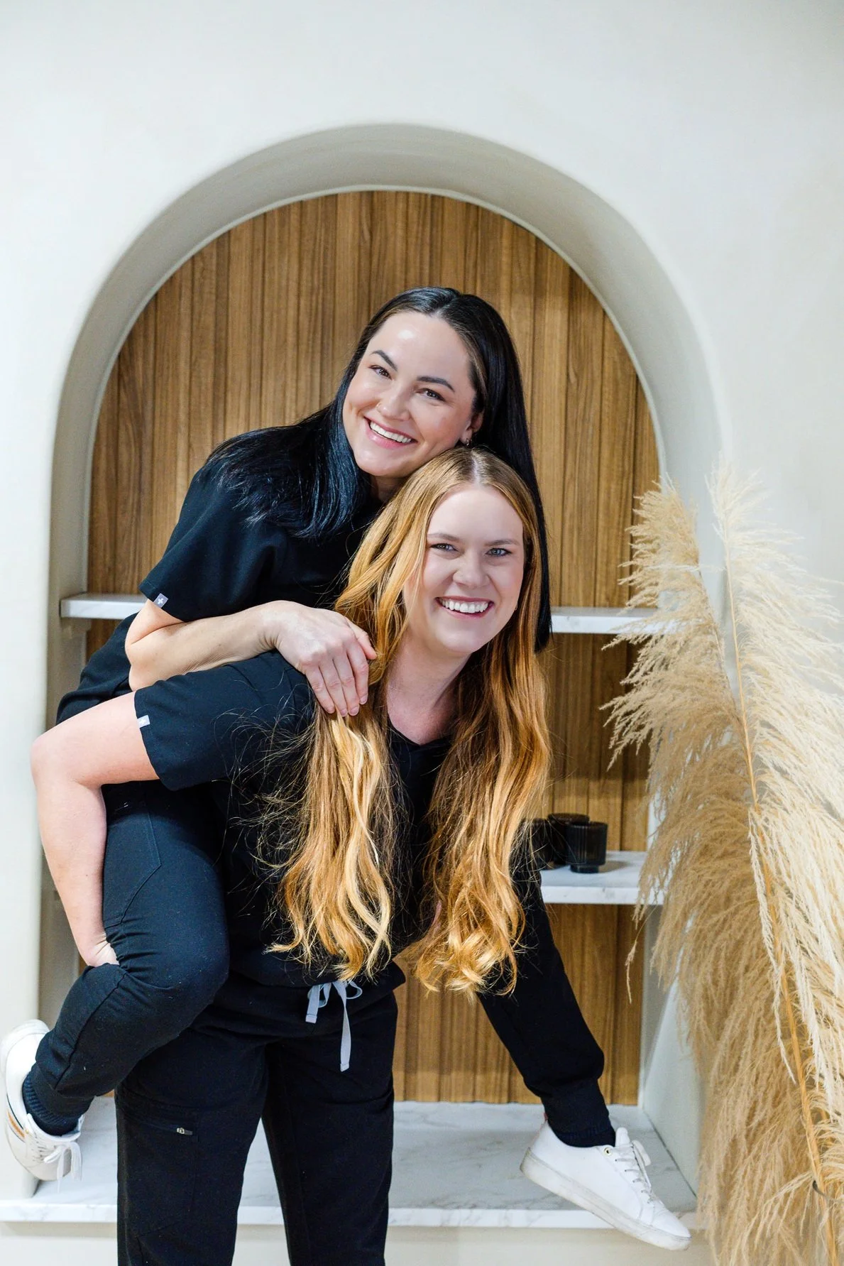 Two women, one in a black outfit with black hair and the other in a black outfit with long red hair, smiling and posing together indoors, with a wooden arch and pampas grass in the background.