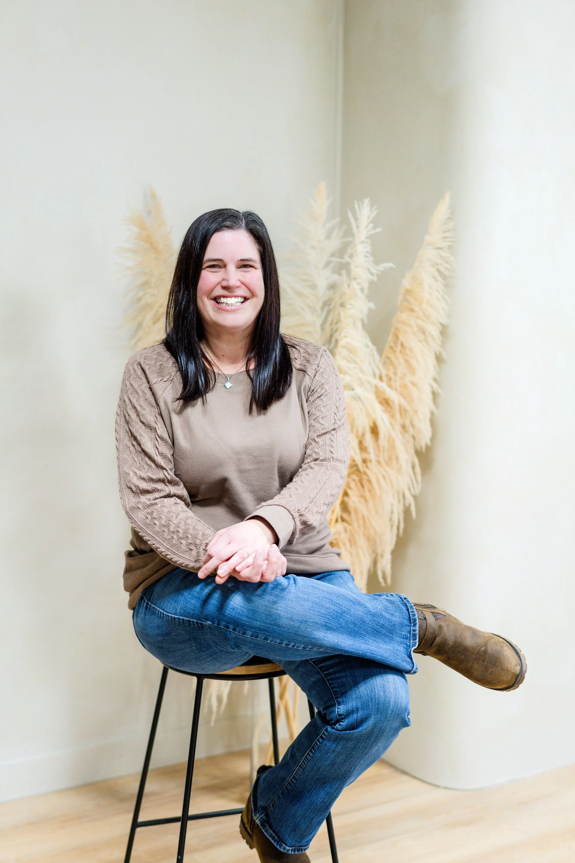 A woman with black hair, smiling and sitting on a stool with crossed legs. She is wearing a beige sweater, blue jeans, and brown boots. Behind her are cream-colored pampas grass decorations in a corner of a room with light-colored walls and wooden flooring.