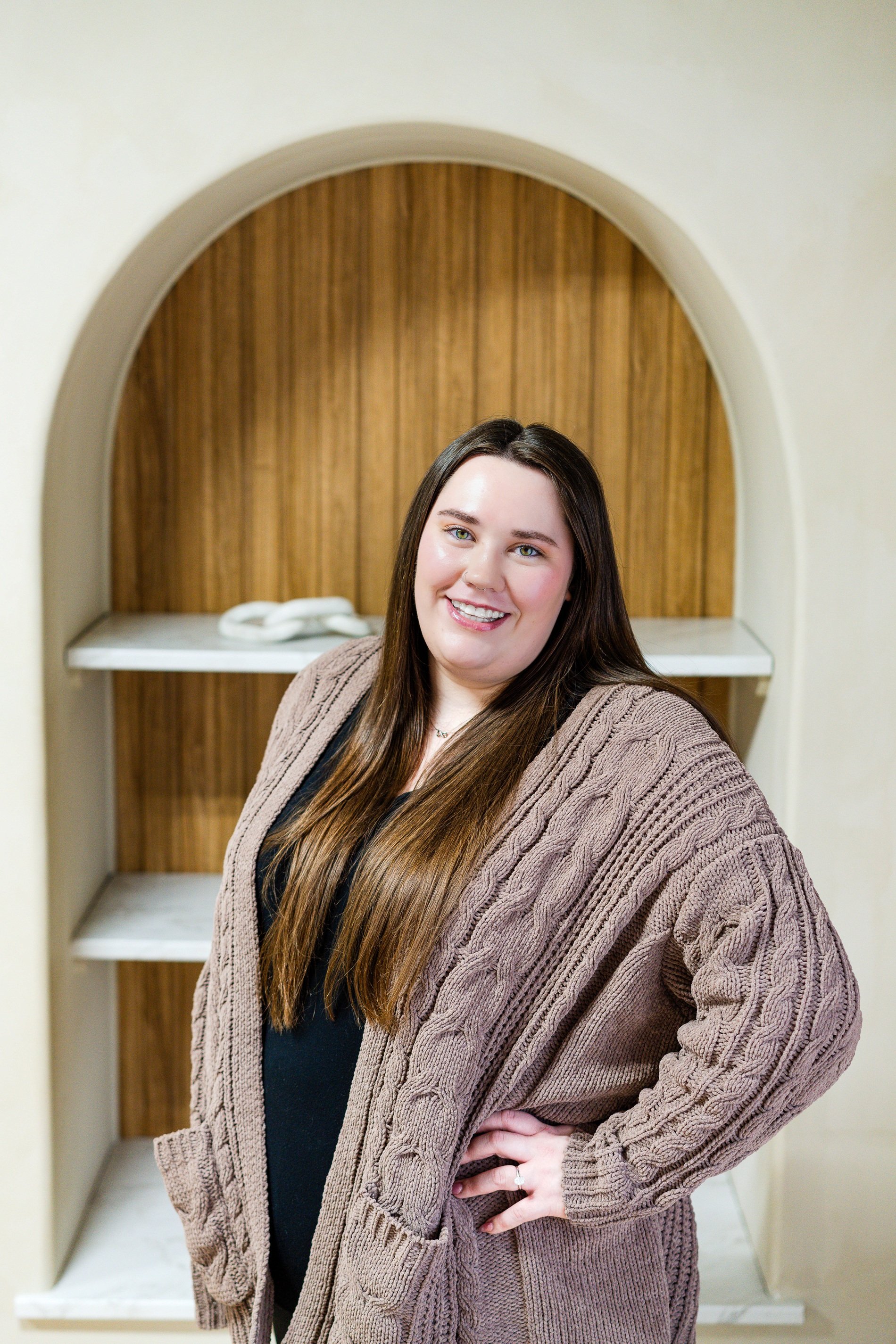 A woman with long brown hair and blue eyes smiling, wearing a black top and a brown knitted cardigan, standing in front of a wooden arched niche.