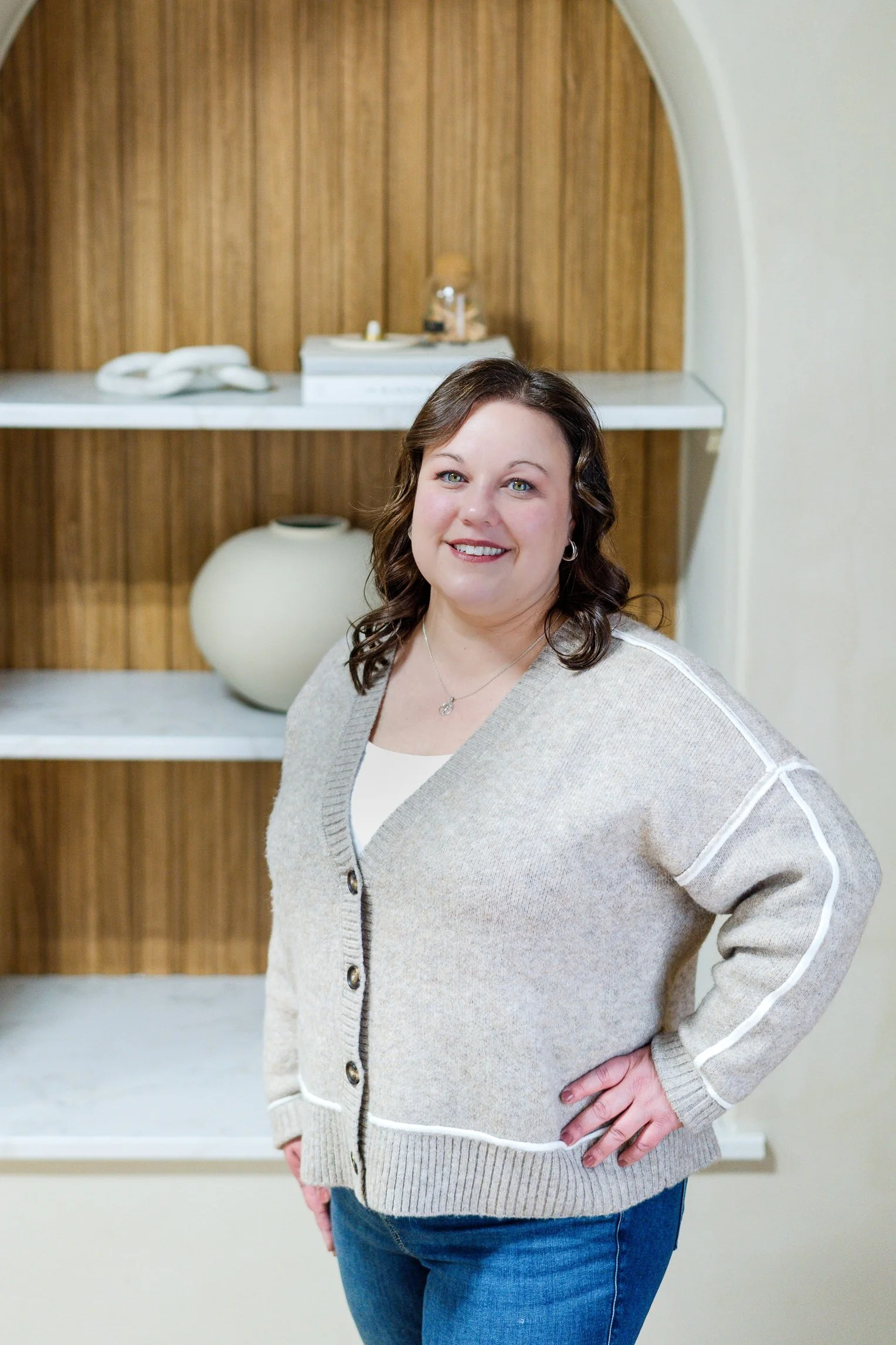 A woman with shoulder-length dark hair, wearing a beige cardigan with white trim and blue jeans, standing in front of a wooden wall with shelves.