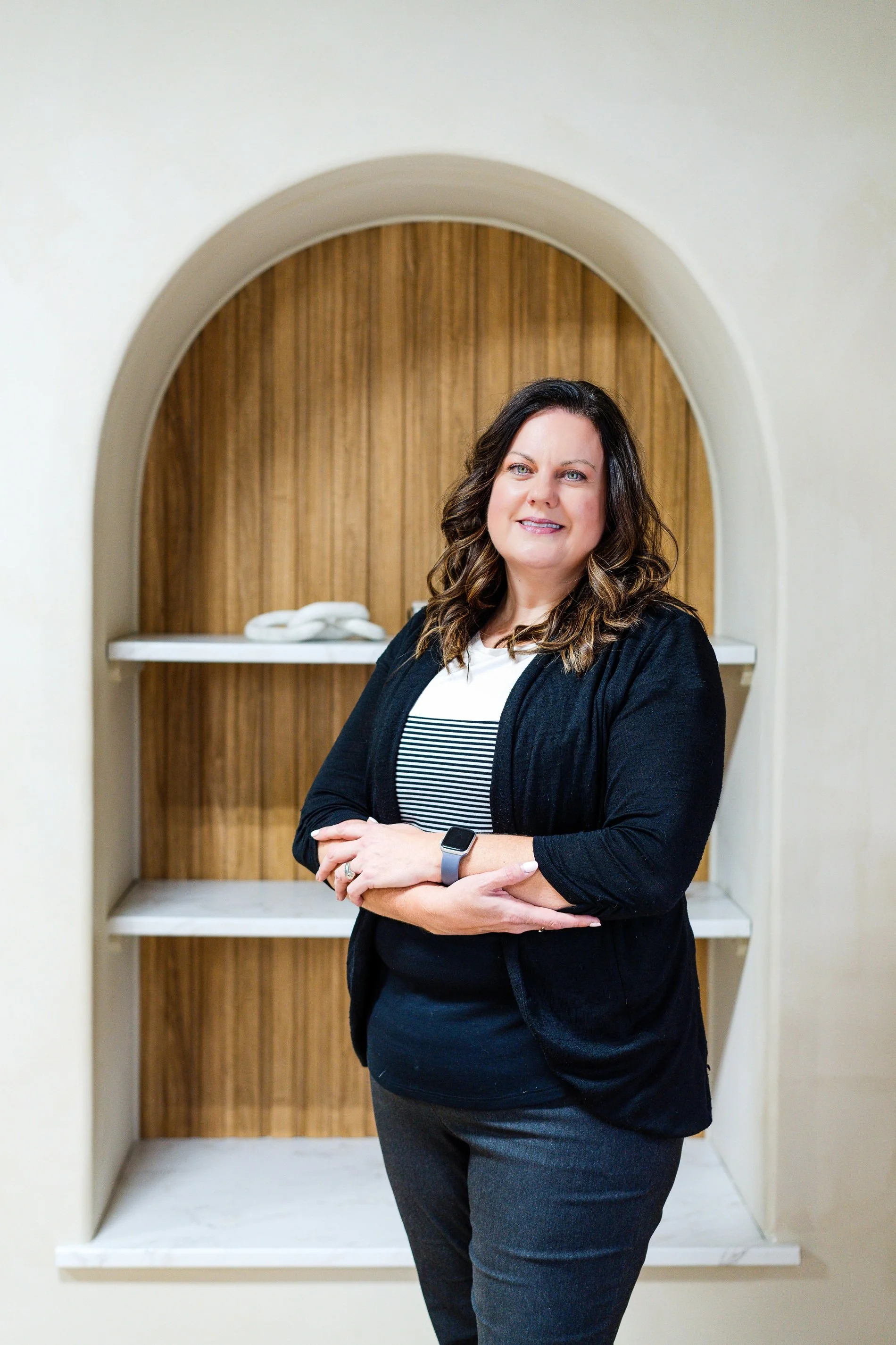 A woman with shoulder-length dark hair, wearing a black cardigan, striped shirt, and smartwatch, standing in front of a wooden arched wall with white shelves and towels.