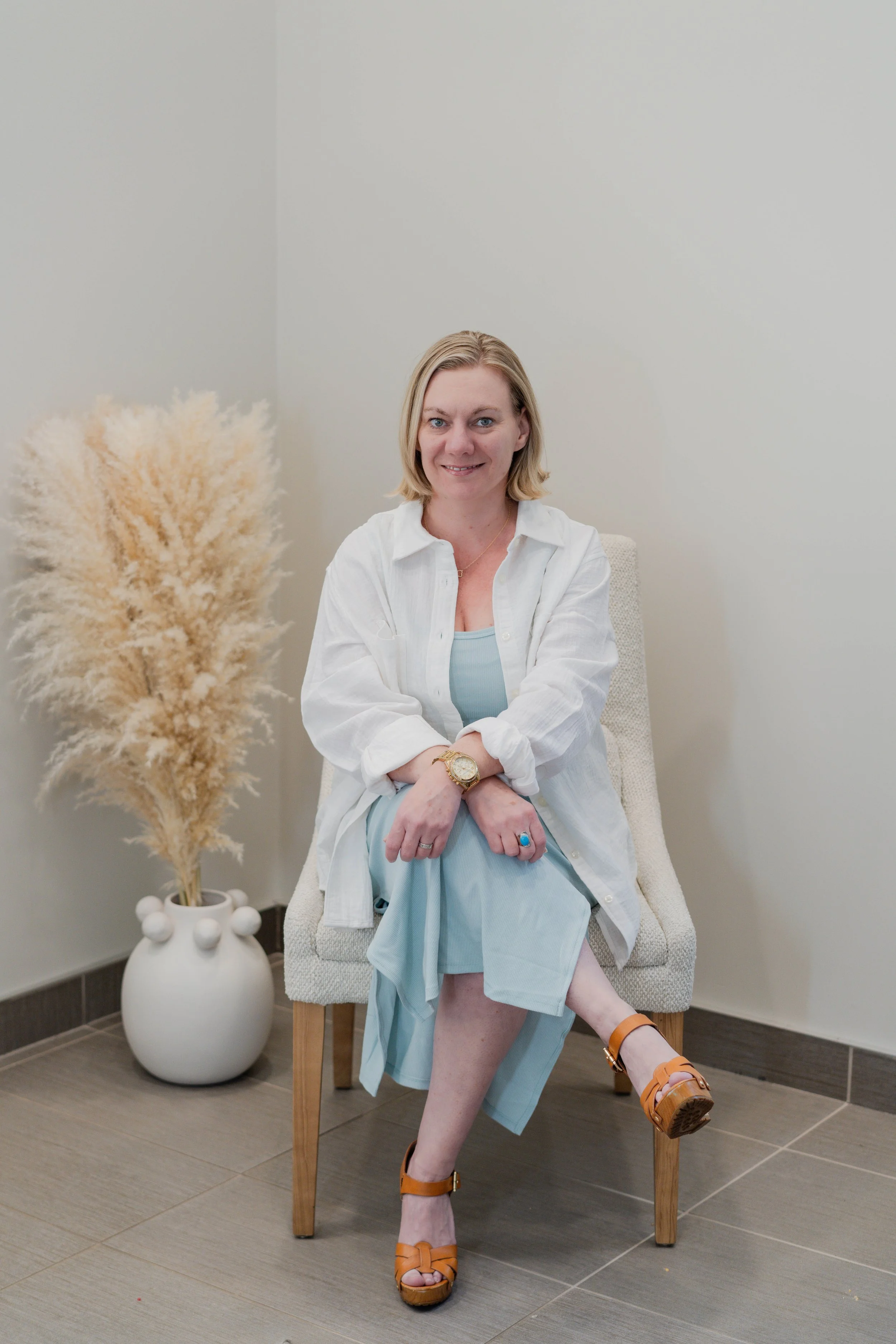 A woman, smiling and sitting with her legs crossed on a cream-colored upholstered chair with wooden legs. She has shoulder-length blonde hair and is wearing a light blue dress, an open white shirt, tan platform sandals, and a gold-toned wristwatch.