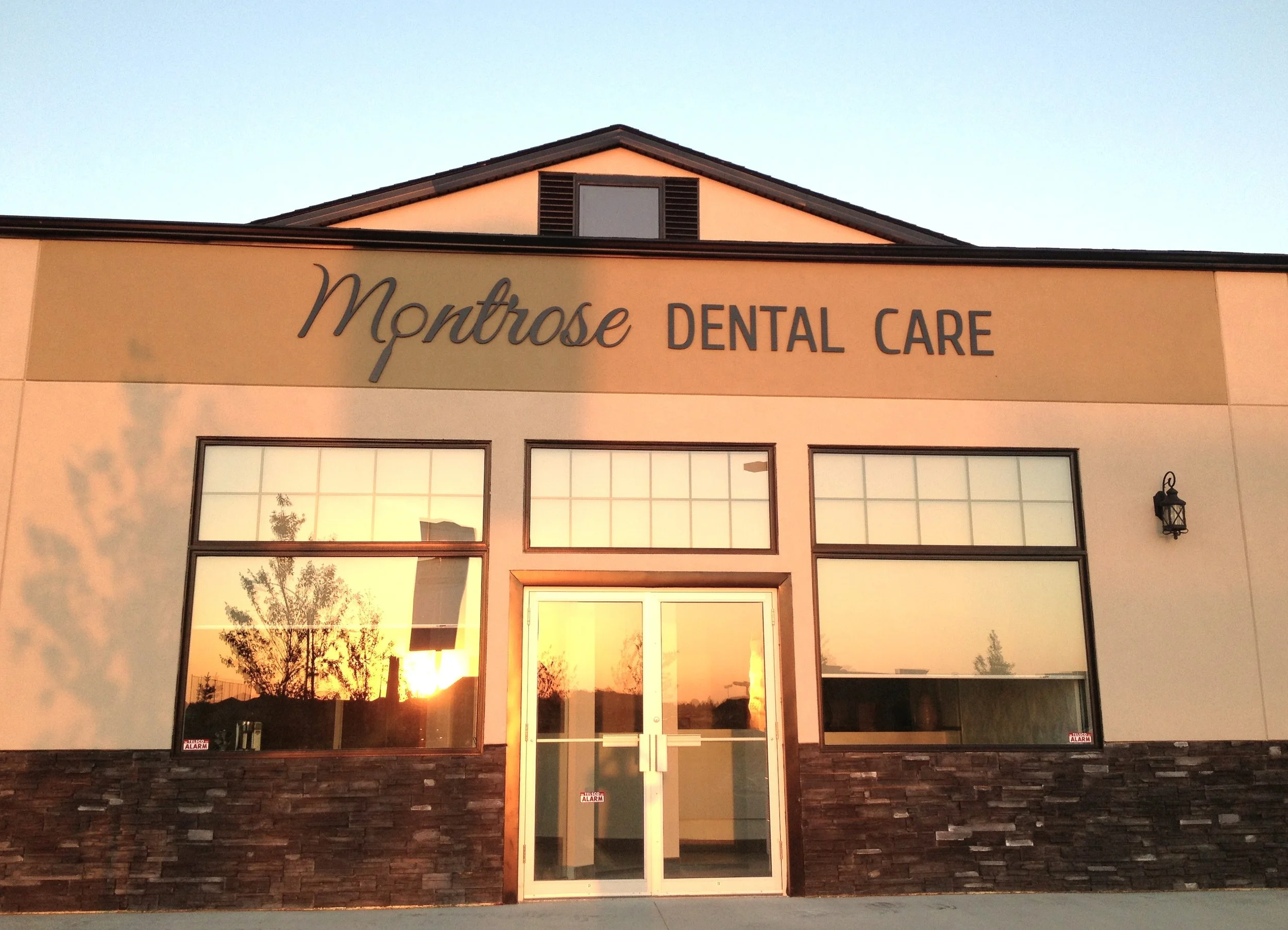 The exterior of Montrose Dental Care building during sunset, with large windows reflecting trees and the orange sky, and a double glass door at the entrance.