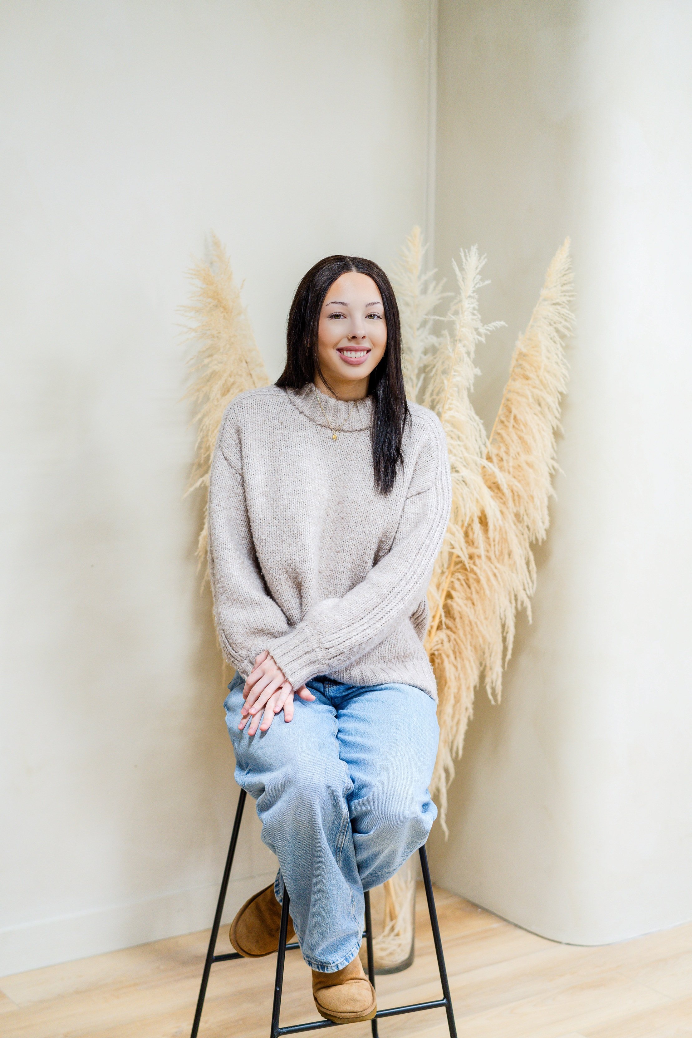 A woman with long wavy brown hair, wearing a black top, gray cardigan, and blue jeans, sitting on a black stool in front of a beige wall with tall, cream-colored pampas grass.