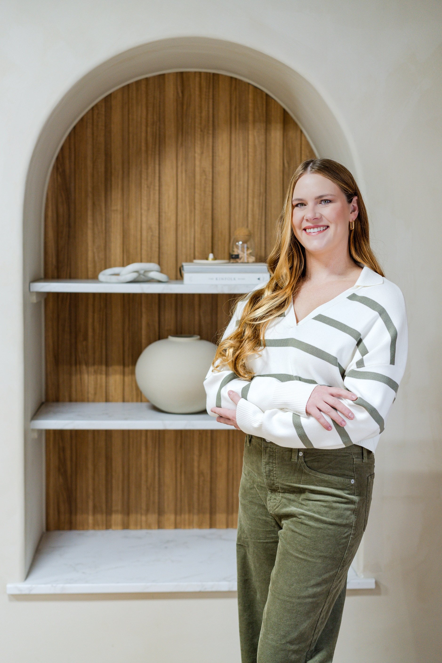 A young woman with long, wavy red hair and a bright smile is standing in front of a wooden shelf with decorative items, wearing a white sweater with green stripes and olive green pants.