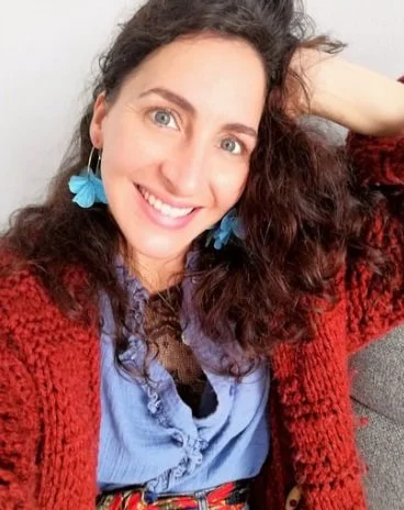 A woman with curly brown hair and blue eyes smiling, wearing blue earrings shaped like butterflies, a blue top, and a red cardigan, sitting on a gray couch with a white wall background.