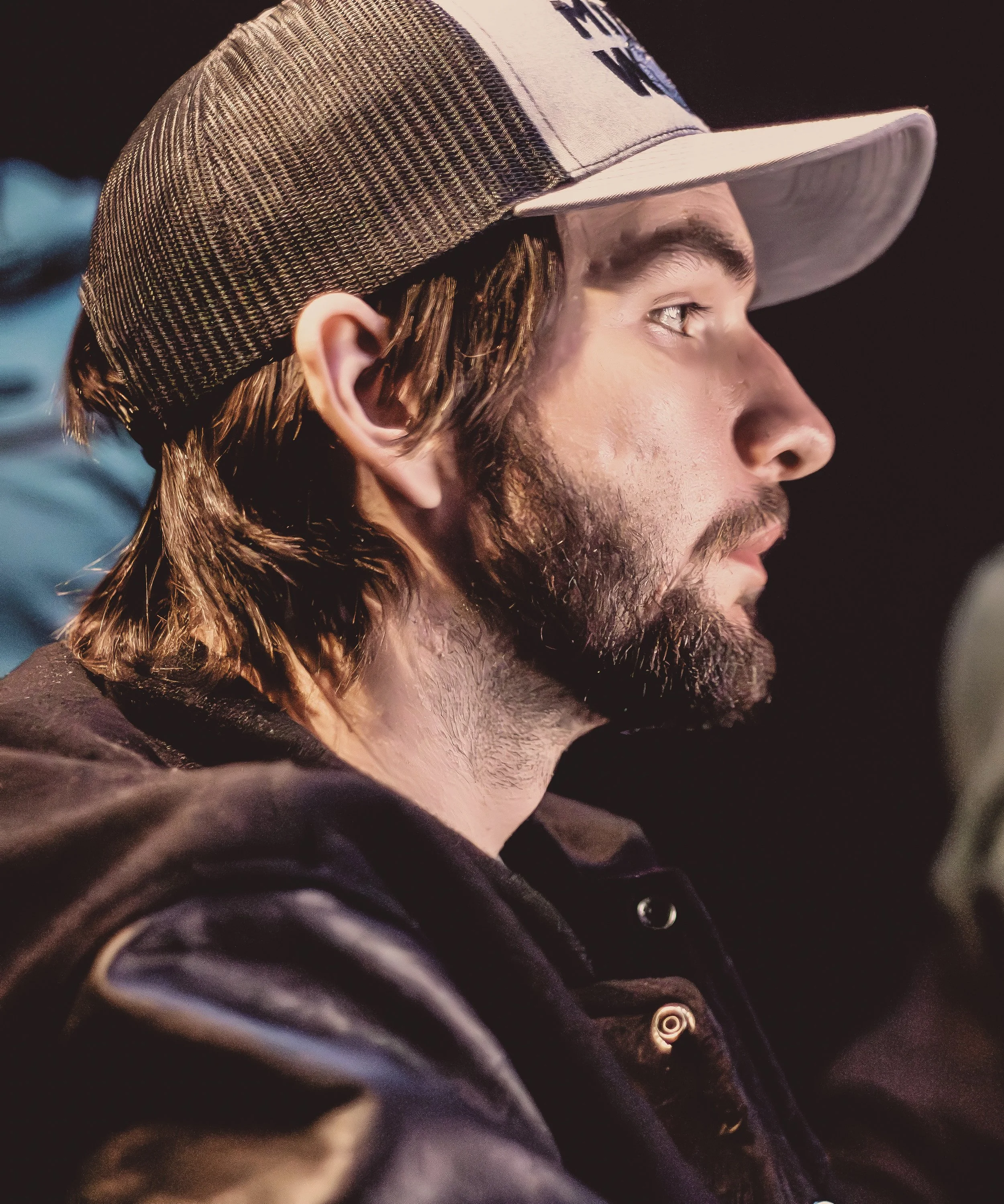 Close-up profile of a man wearing a baseball cap with textured fabric and a jacket with metallic buttons, against a dark background.