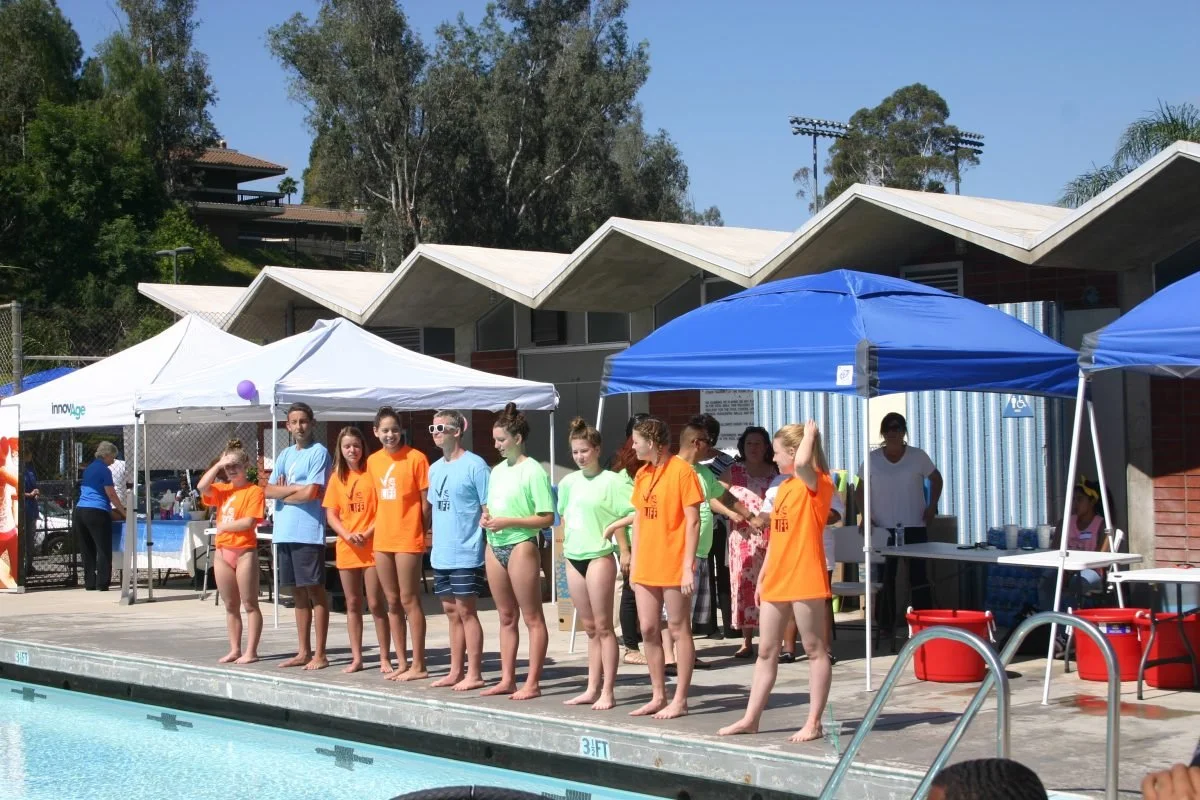 Swimmers in colorful t-shirts lined up at the edge of a swimming pool for a competition or event, with tents and a building in the background.