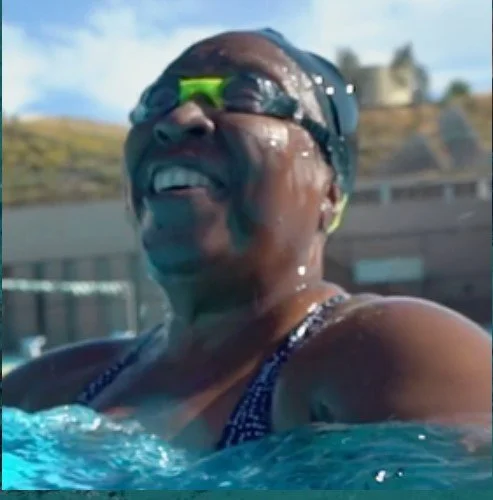 A woman wearing swim goggles and a swimsuit smiling in a swimming pool.