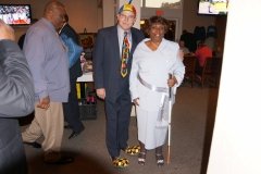 A group of people standing together in a social setting, including a man in a dark suit with a patterned tie and a woman in a white outfit. They appear to be engaged in conversation.