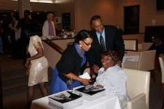 A woman wearing glasses and a blue dress signing something at a table with a young girl in a white dress, while a man in a suit looks on. In the background, other people are mingling.