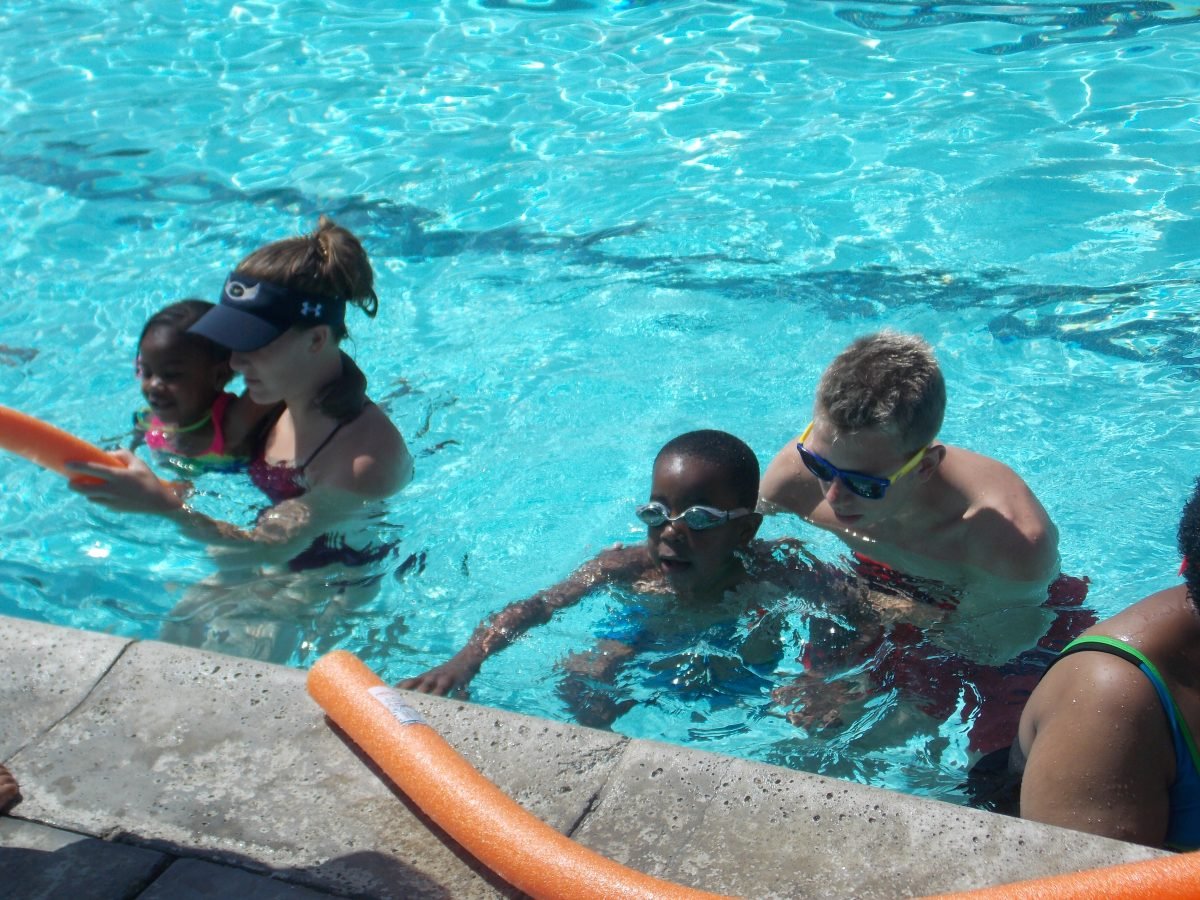 Children and adults in a swimming pool, some wearing goggles, playing and swimming.
