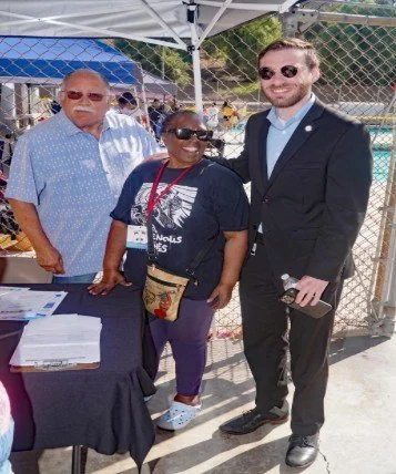Three people smiling at an outdoor event near a chain-link fence, with a table covered with documents in front of them.