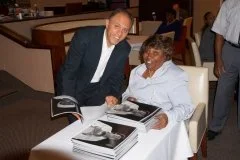 A man in a dark suit and white shirt, and an older woman in a white blouse, sitting at a table with several copies of a book, smiling and looking at the camera.