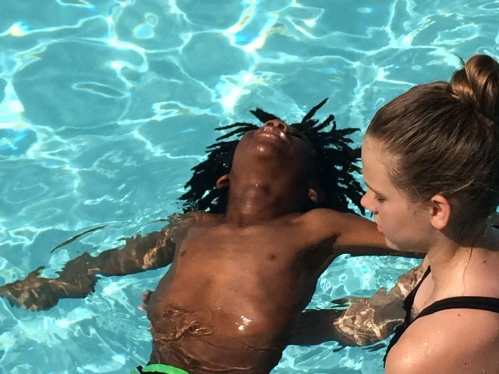 A woman assists a man with dark braided hair and dark skin in a swimming pool, holding his shoulders as he floats with his eyes closed.