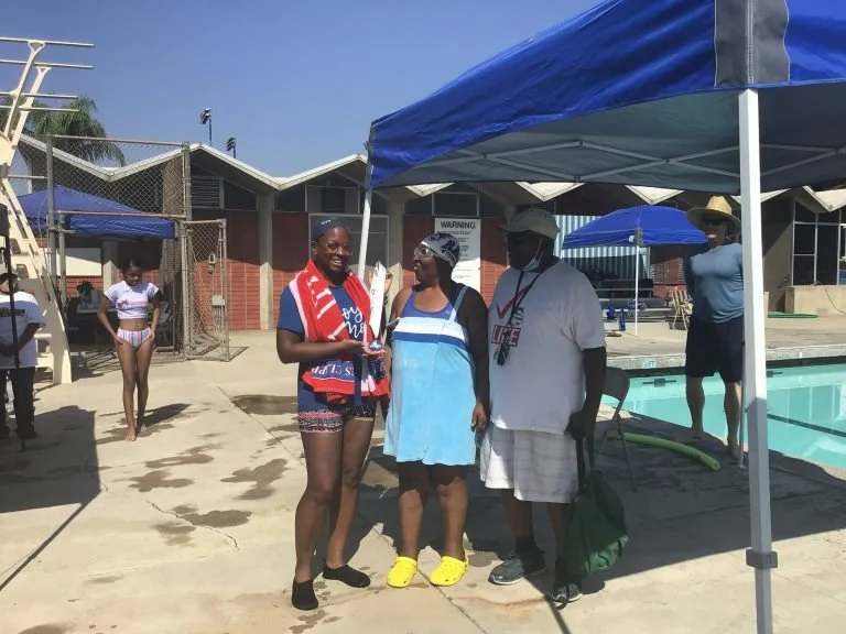 Three people standing under a blue canopy near a swimming pool, smiling and talking. Two women and one man, with people in swimsuits and poolside equipment around.