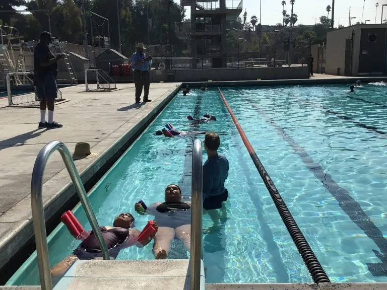 Swimmers lying on floaties in a swimming pool, with an instructor standing nearby. Two people are on the pool deck, one holding a clipboard and another taking photos or videos. The pool has lane dividers, and there are people swimming in the backgrou