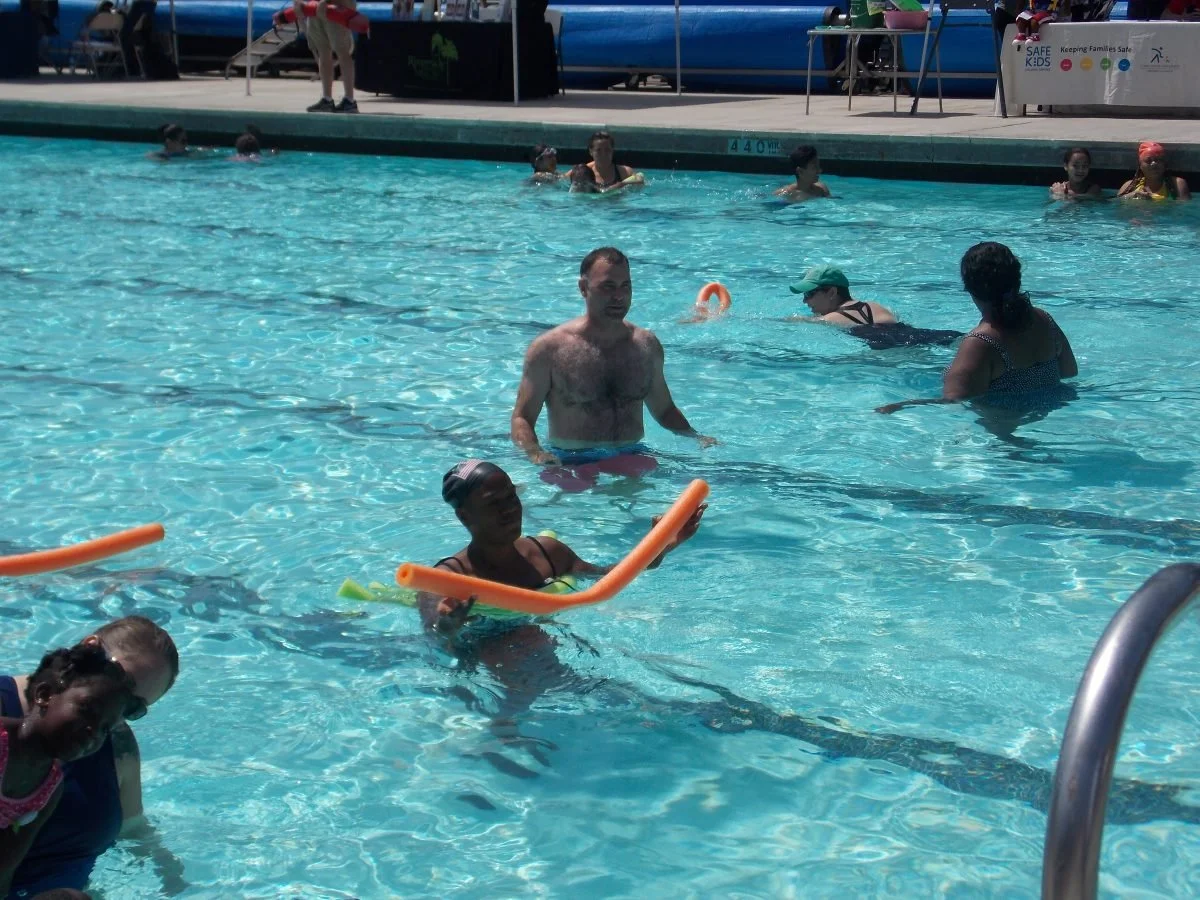 People participating in a swim class or water activity at an outdoor pool, with instructors and children using pool noodles for safety and support.